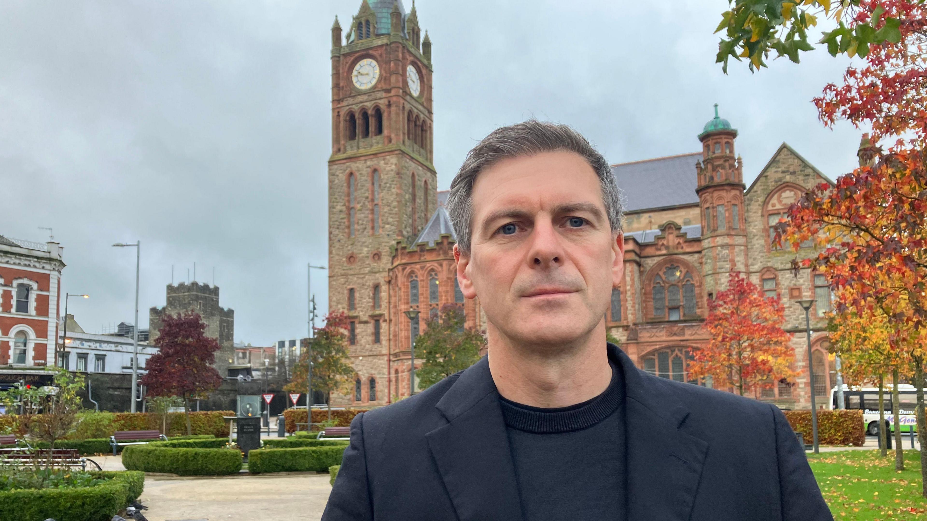 Councillor Shaun Harkin, in a black jacket and sweater, stands in Derry's pece garden. A number of hedges planted in a paved park can be seen in the background as can the city's Guildhall