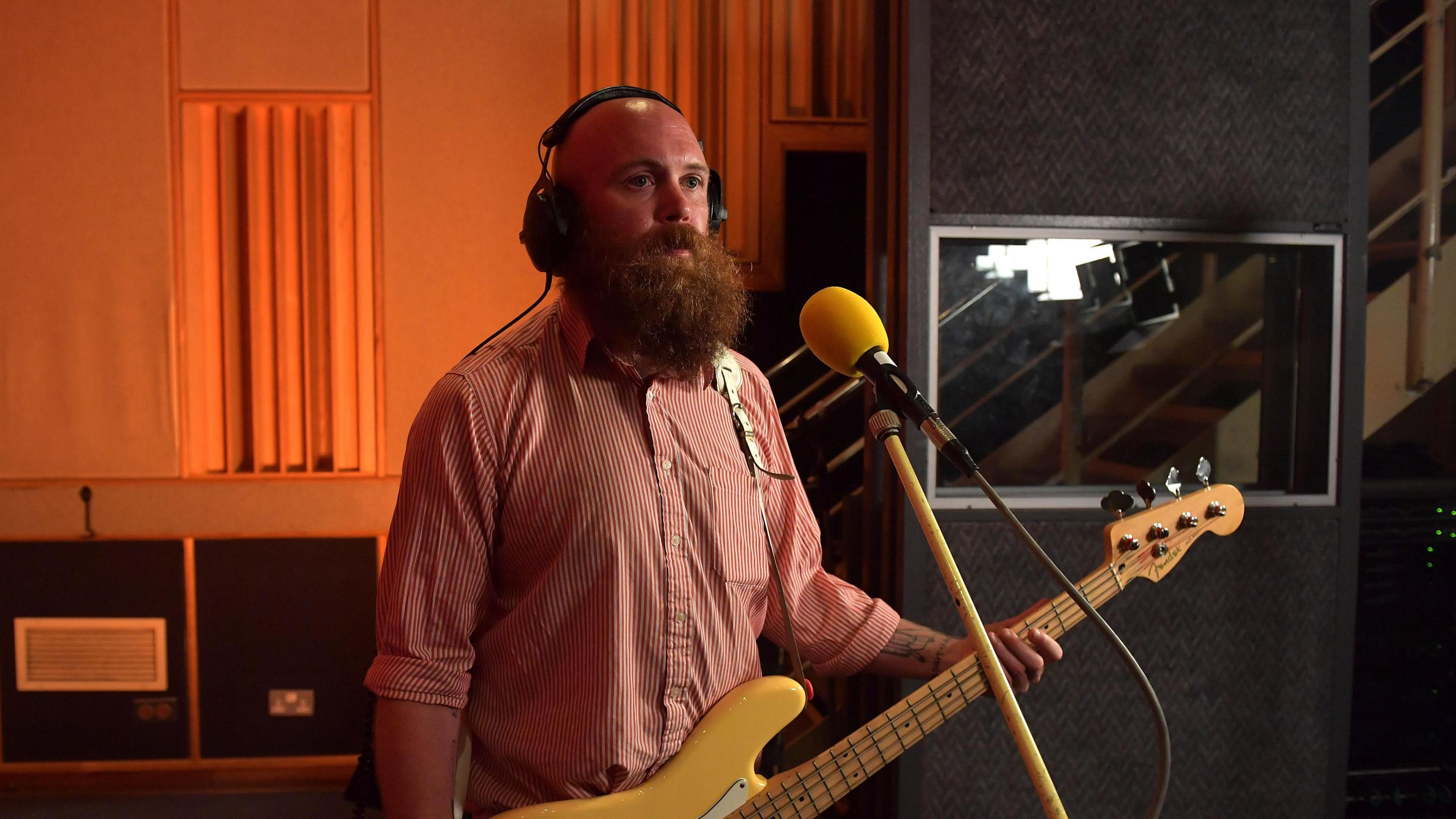 Adam Devonshire is holding a bass and standing in front of a microphone. He is wearing a red shirt and headphones. He has a ginger beard.