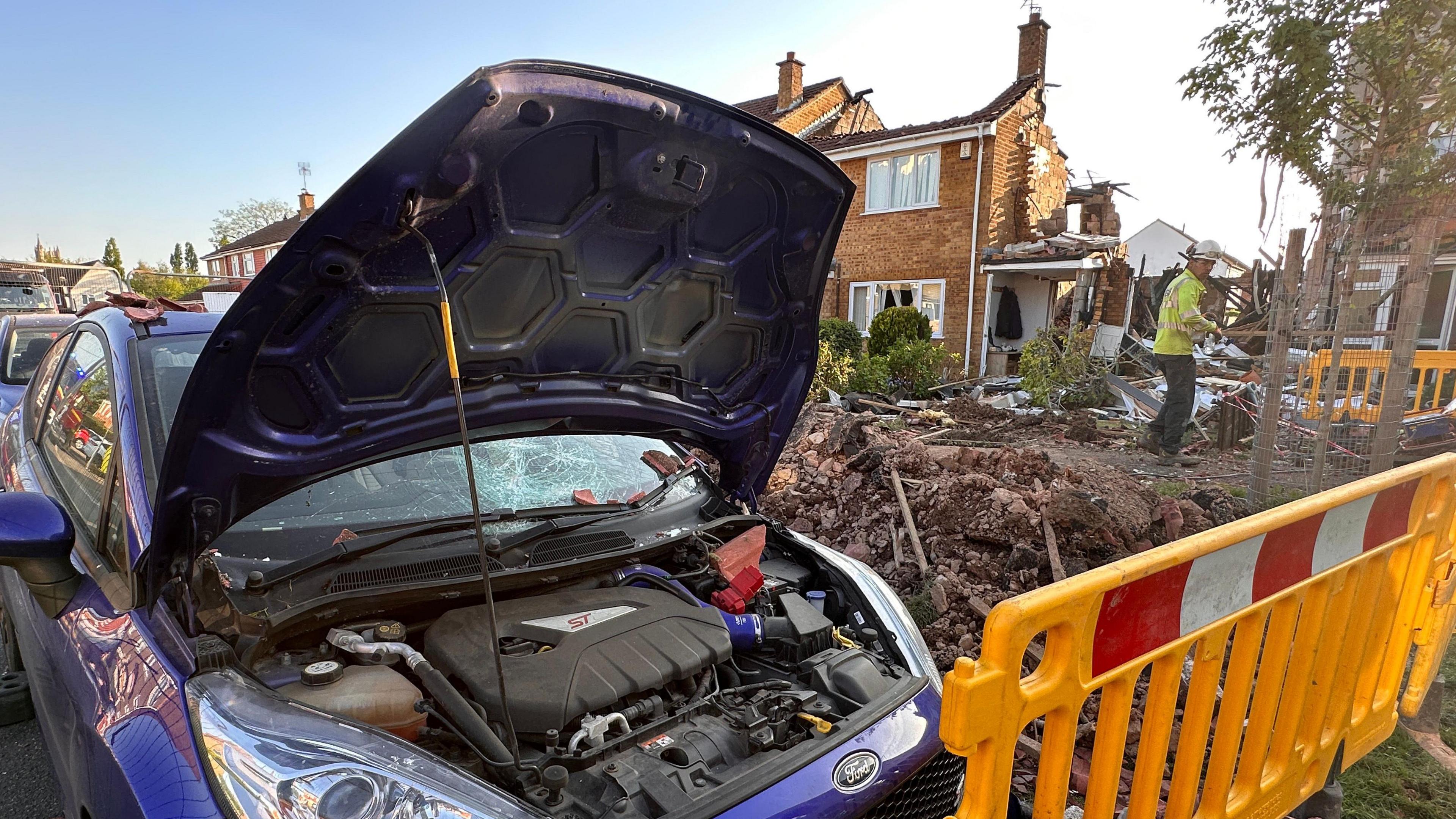 A damaged blue Ford parked on the street in front of the property where a suspected gas explosion happened. The bonnet is up on the car and the windscreen is smashed. Debris can be seen next to the car and the damaged house is in the background. A yellow gate is cordoning off access to the house.