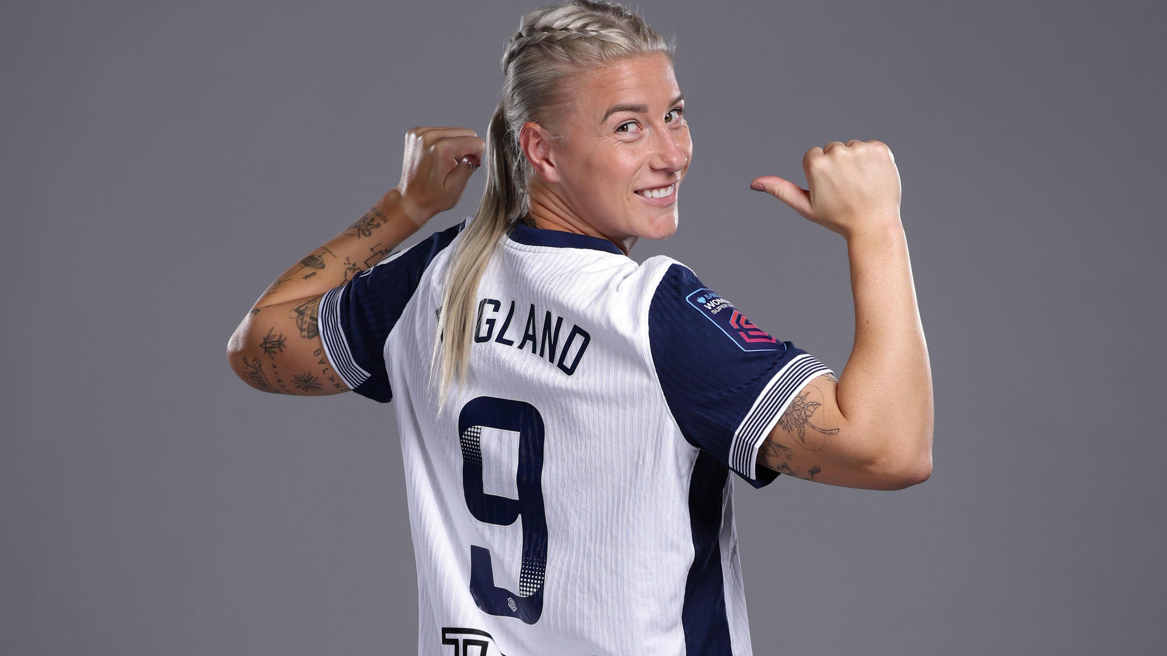 Bethany England in a blue and white England jersey. She has her hair in plaits and points at the back of her shirt.