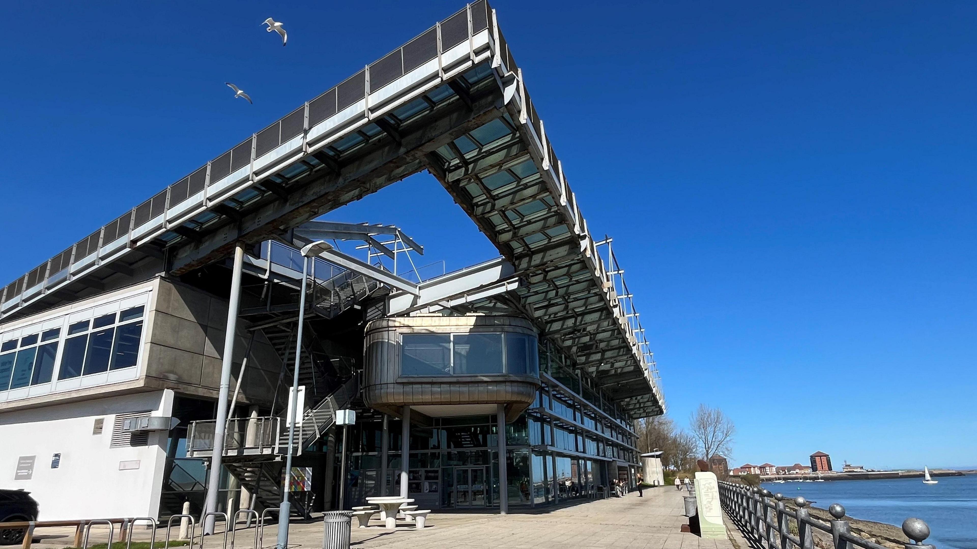 The National Glass Centre is a tall building made of metal panels and glass, overlooking the river Wear. There is seating space and a cafe in front of it, with a dog walker in the distance.
