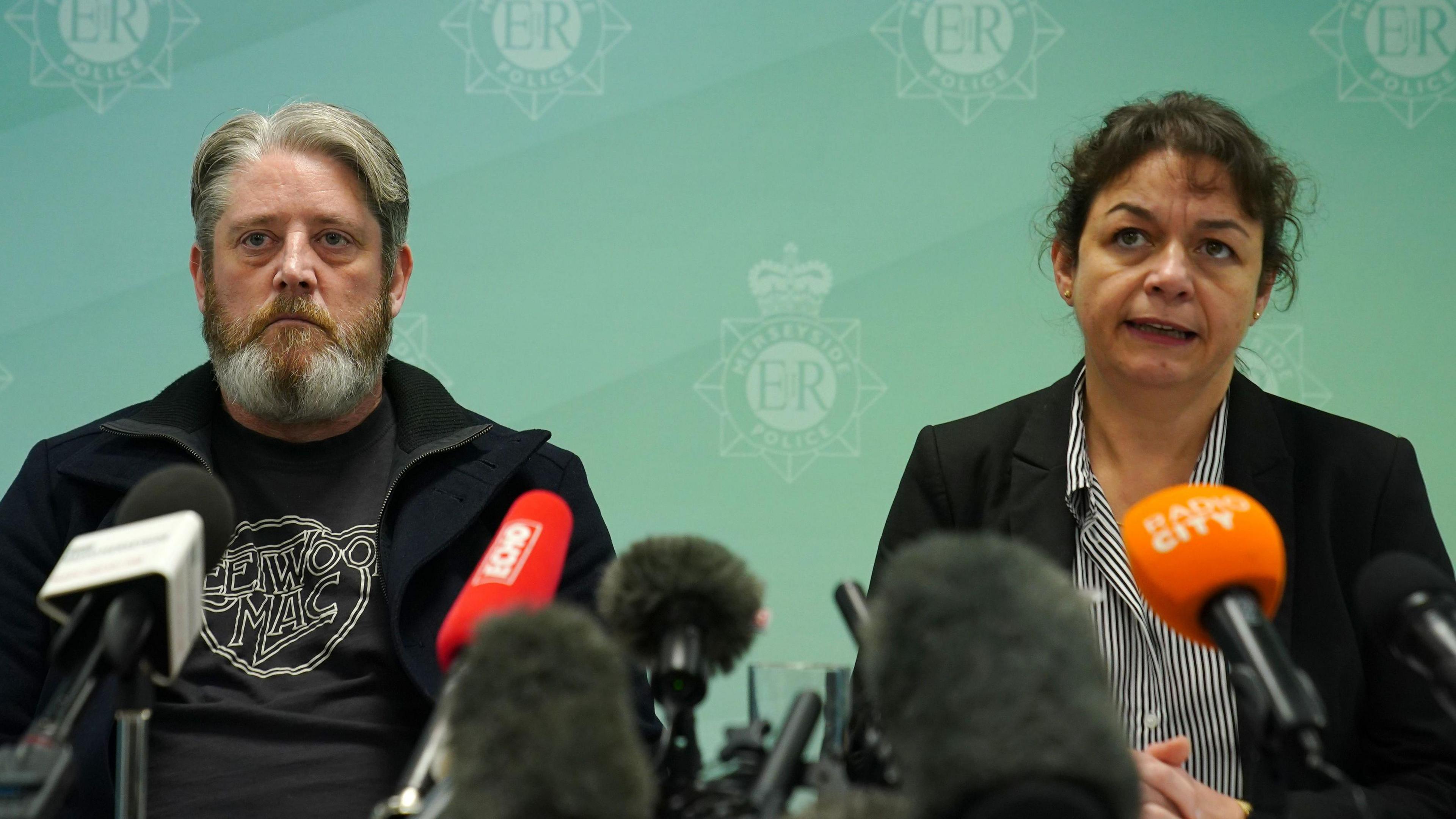 A sombre looking Tim Edwards, with grey hair and a grey beard, sitting in front of a series of microphones alongside Det Supt Sue Coombs, who has black hair tied into a bun and wears a black blazer 