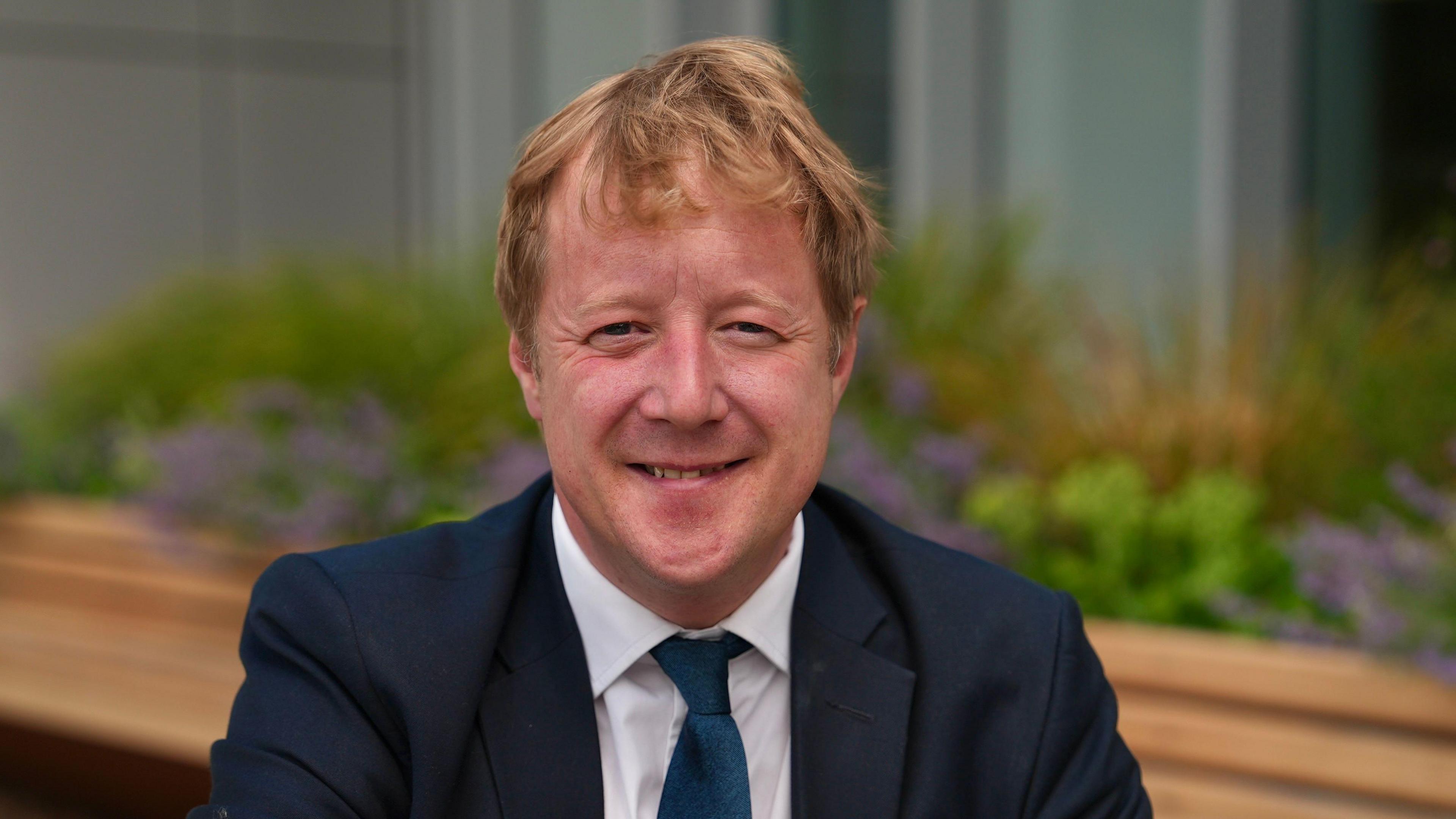Paul Bristow stares at the camera. He wears a dark navy suit jacket, a white shirt and a navy tie. In the background is a beech coloured wooden ledge and a variety of plants.