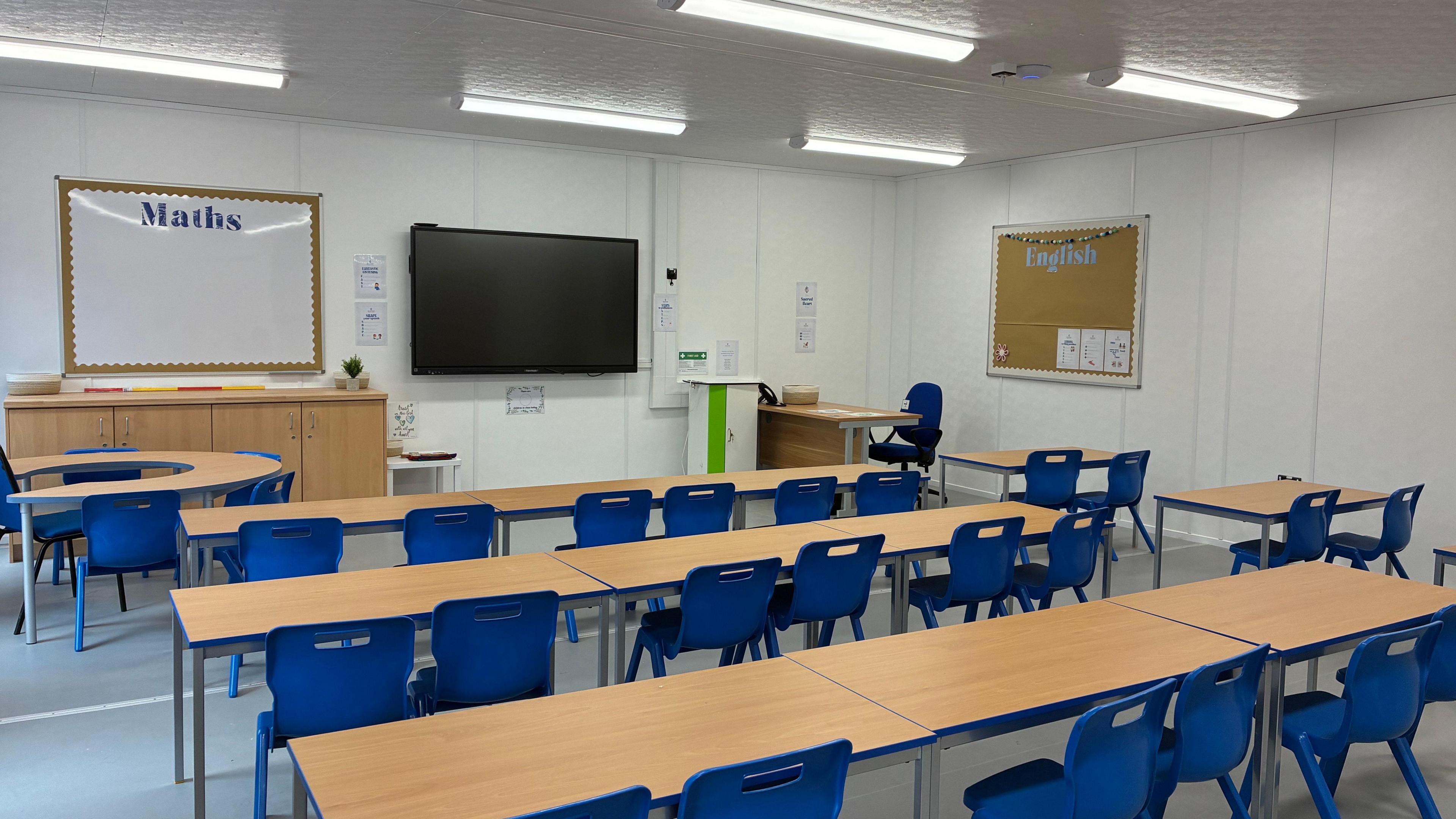 The new temporary modular building in the grounds of St Bernard's Catholic High School. The classroom has wooden desks and blue chairs, with screens and whiteboards on the walls.