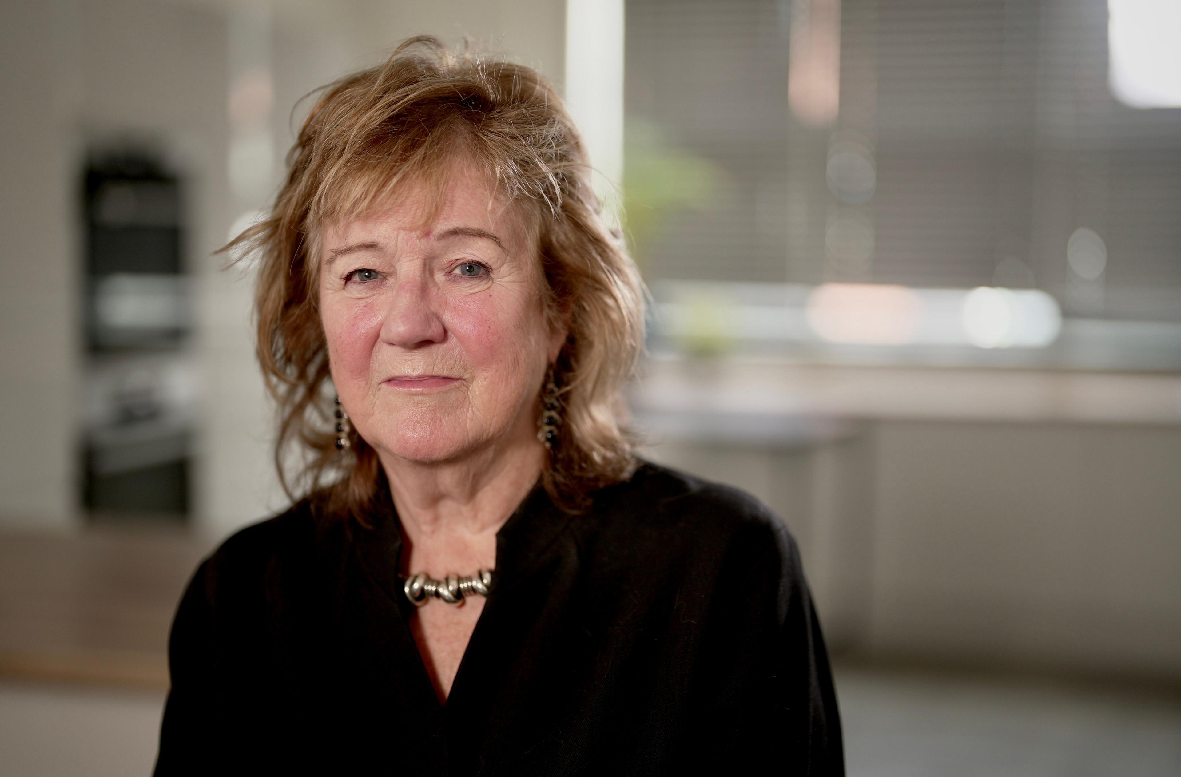 Headshot of Prof Alexis Jay. She has shoulder-length, fair, wavy hair with a fringe, with long dangly earrings with black stones. She is wearing a black collar-less shirt and a chunky silver necklace. She is pictured sitting indoors, with a window and metal blinds behind her.