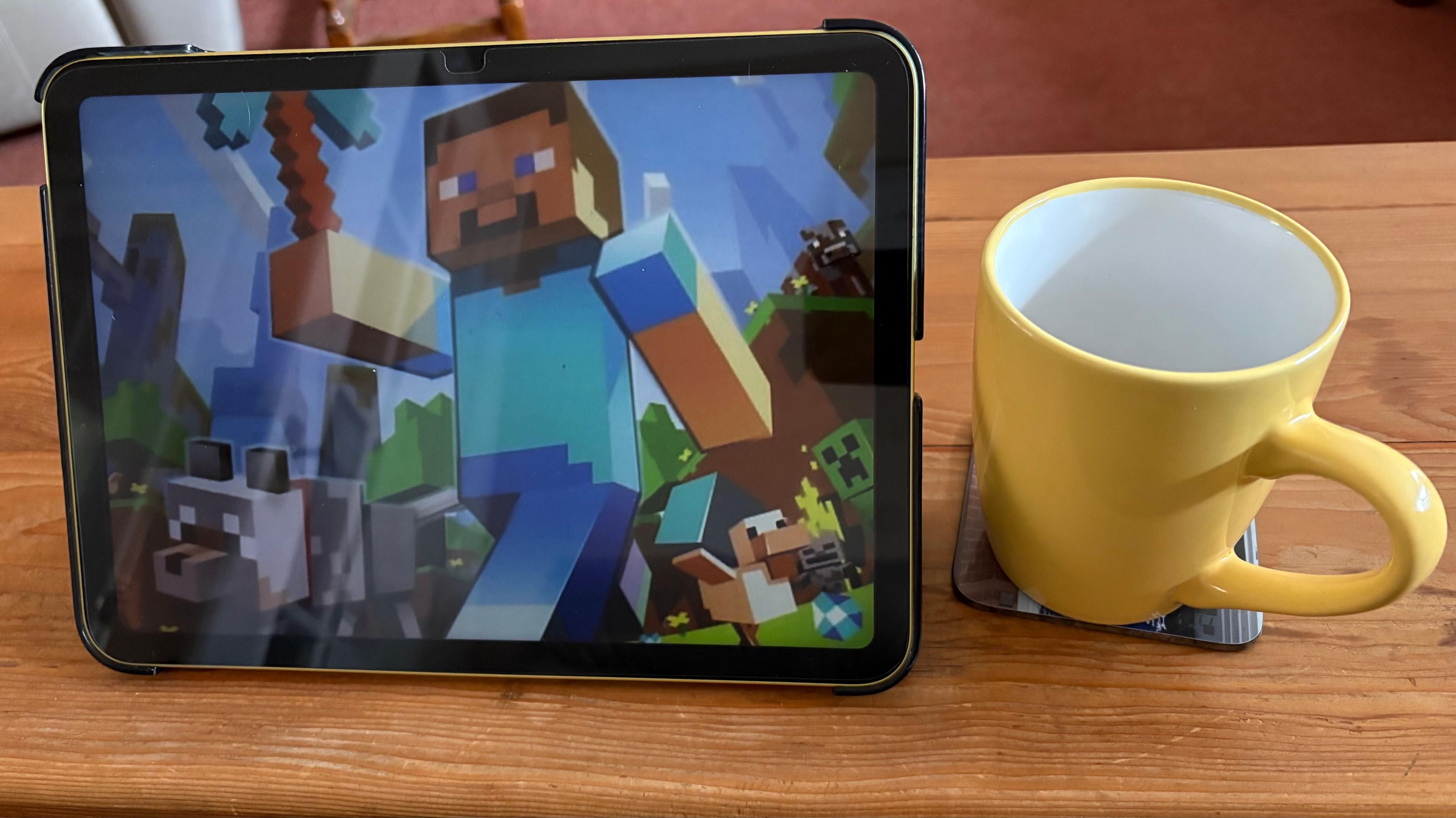 An iPad showing a scene from Minecraft, featuring a character wearing blue clothing fashioned from blocks. The iPad is standing on a table next to a yellow mug.