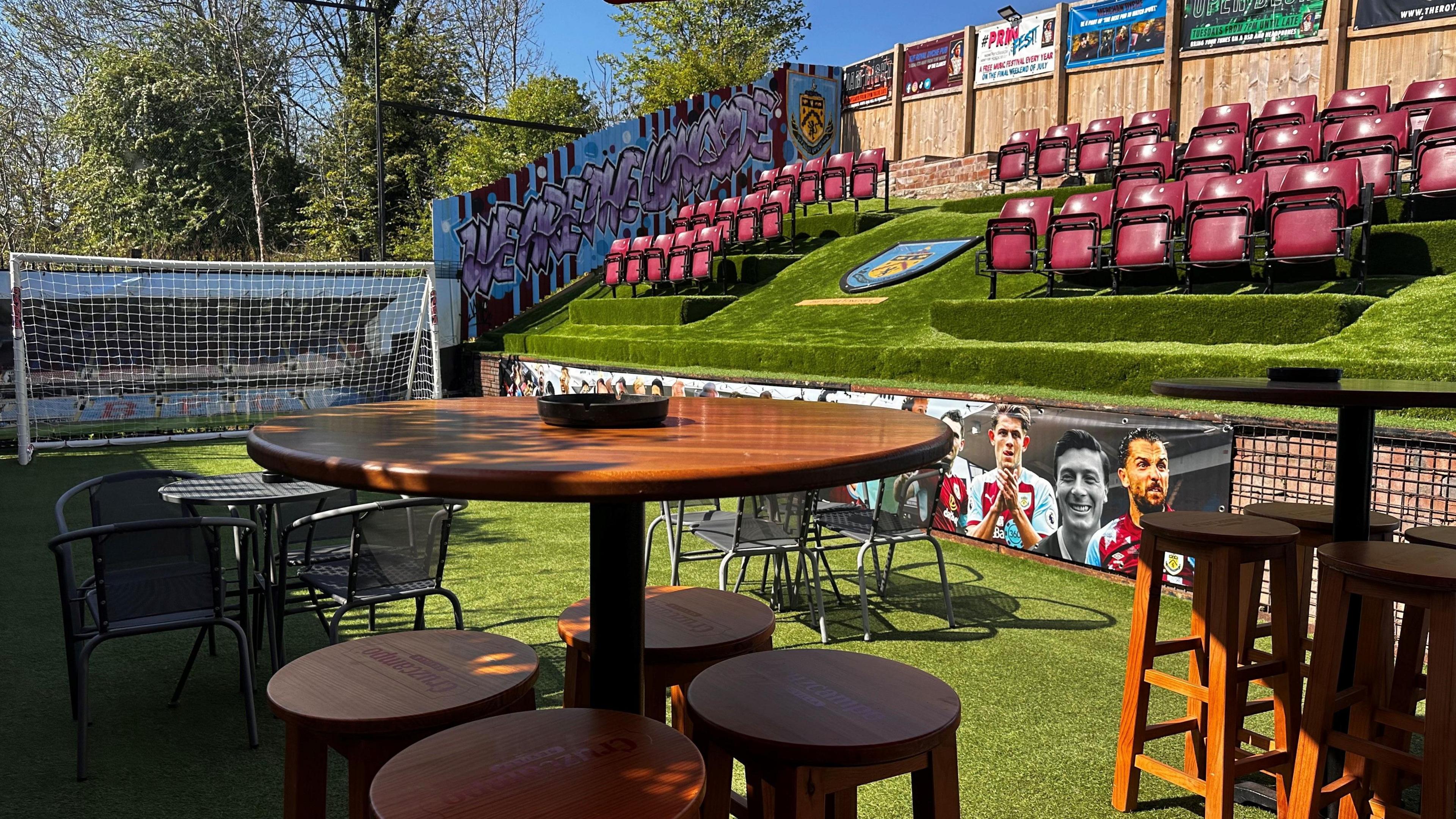 A beer garden transformed into a mini stadium with claret coloured seats like the ones at Burnley's Turf Moor and football net with tables and high stools. It is a sunny day.