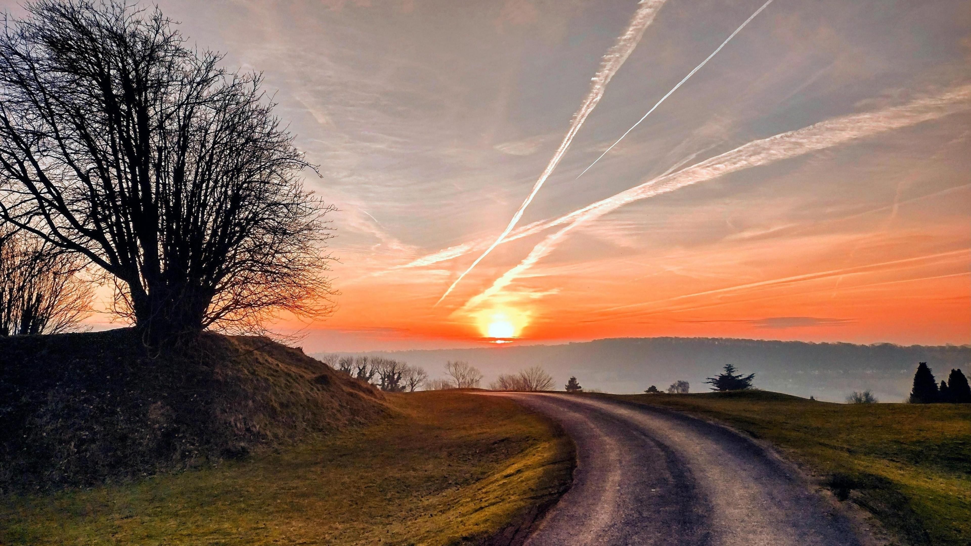 An orange sky and tarmac road leading to a sunrise in the background with trees on a hillside also visible