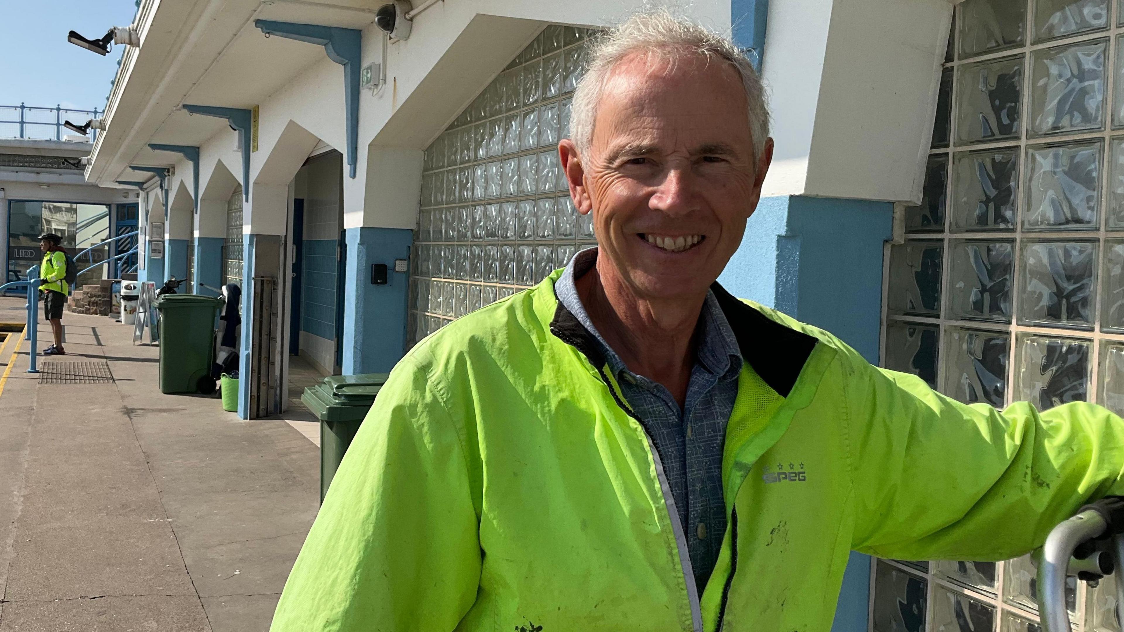 Mike Walker: a man with white hair smiling with teeth, wearing a hi-viz yellow jacket and a blue shirt underneath, behind is glass paned walls and a white and blue building.