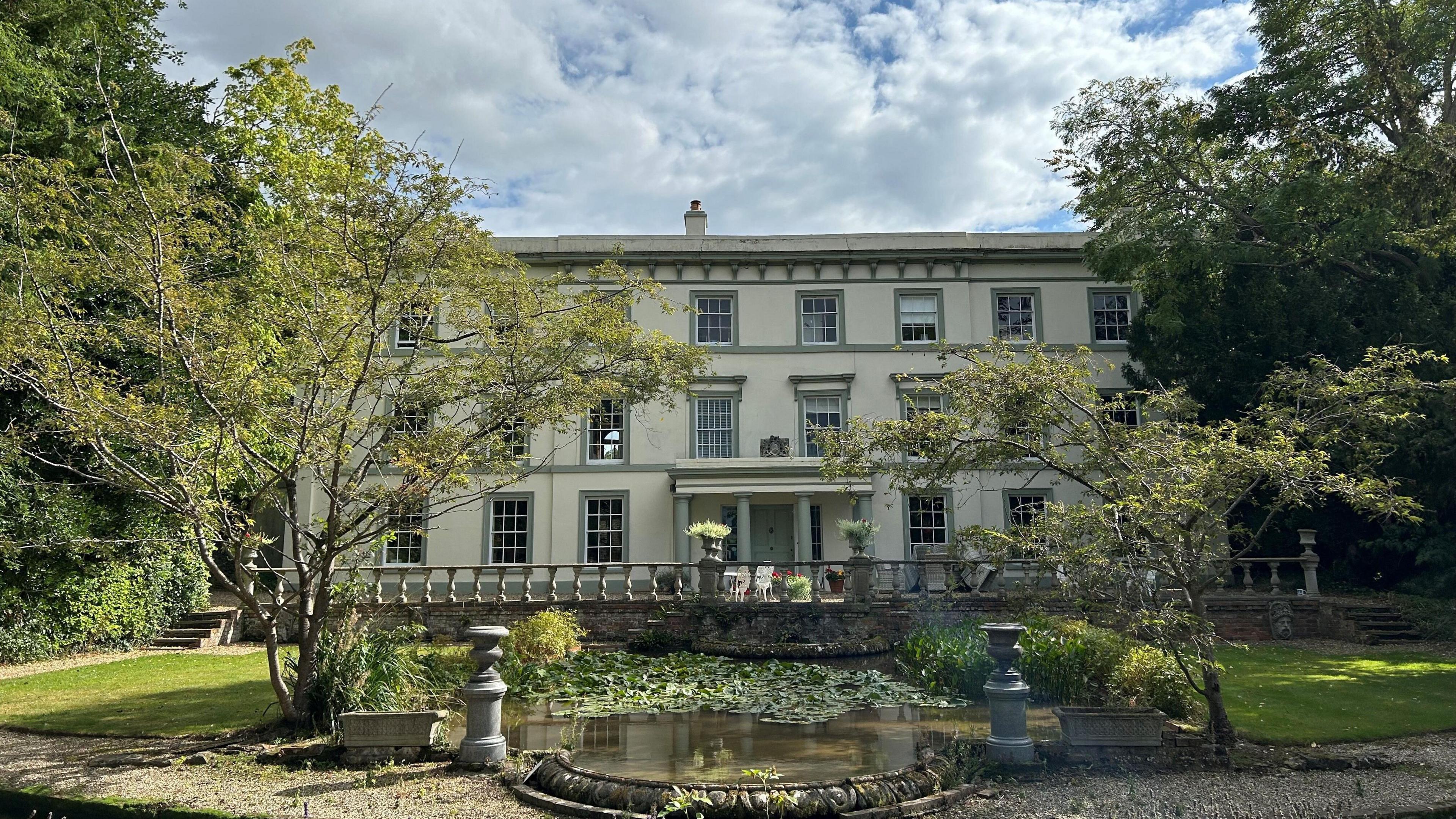 The front of Fyfield Manor. It is a large white building with pastel green trim. It has a pond complete with lily pads in front of it.