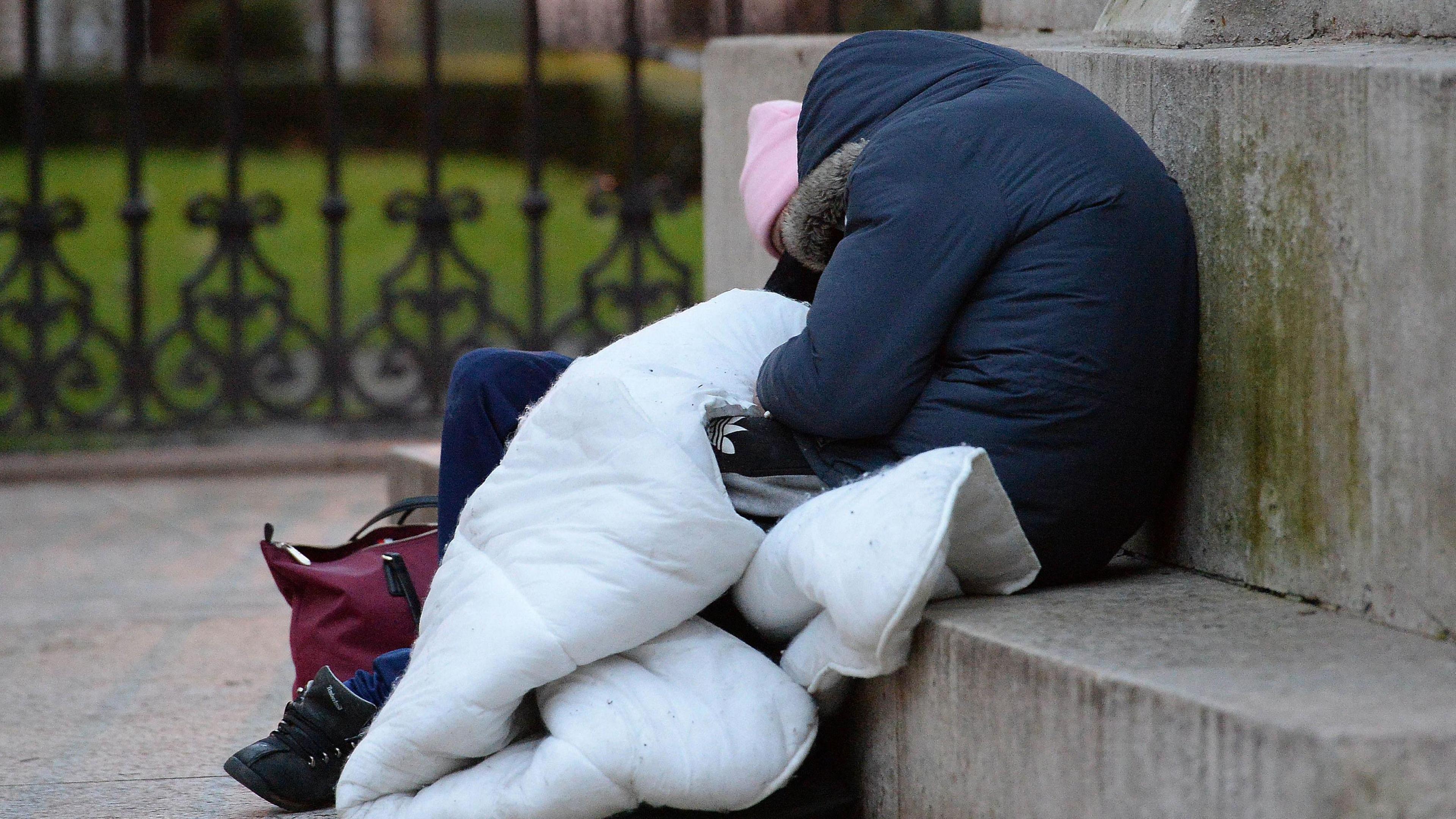 A person is sitting on some steps outside. They are wearing a blue coat and are covered with a duvet. They appear to be sleeping.