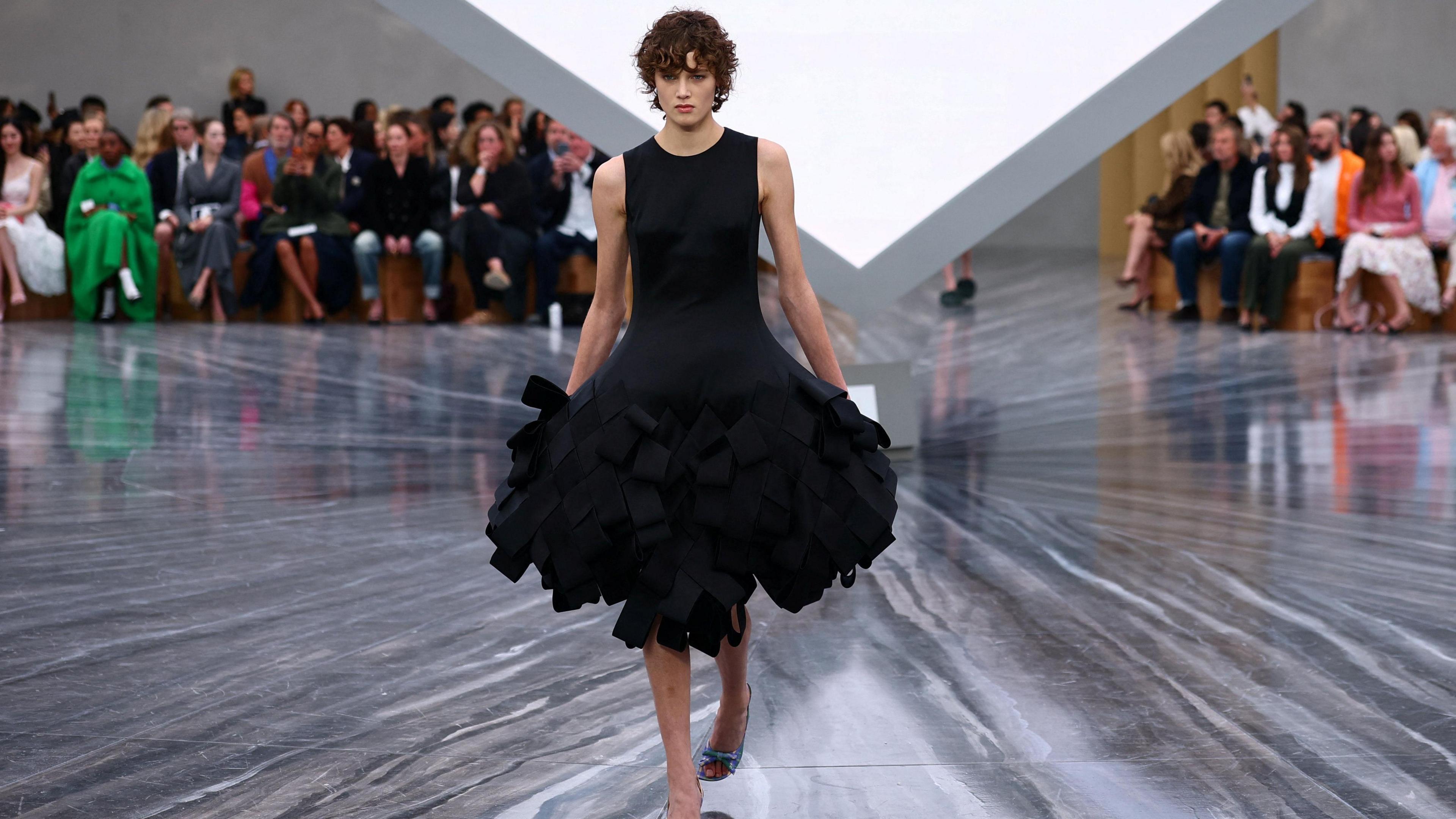 A woman in a black dress with brown curly hair. She is on a catwalk, with the audience behind her.