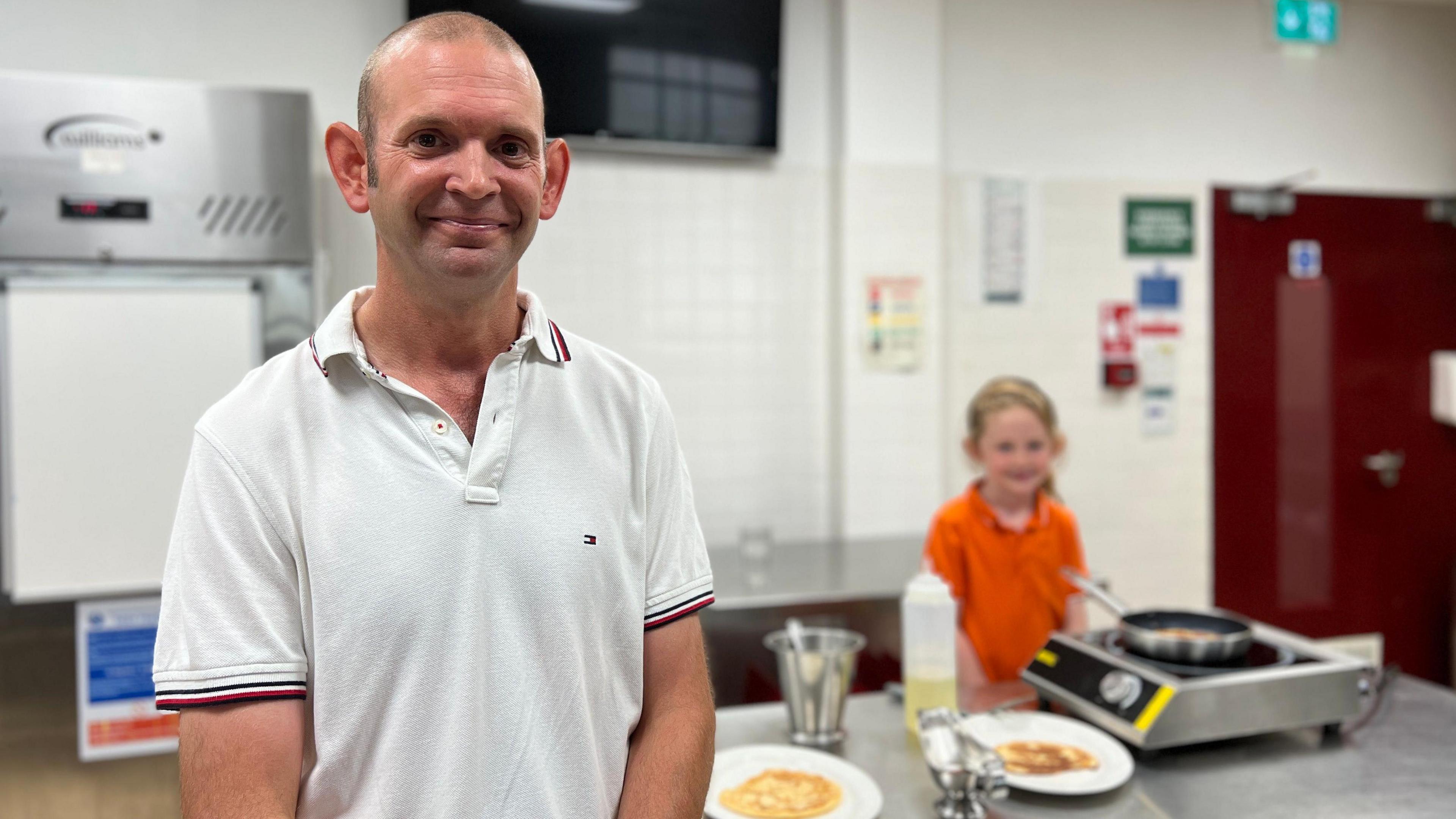 Adrian is in a white polo shirt with red and black trim smiling at the camera in a kitchen setting, with Ophelia in an orange shirt preparing food on a counter behind him. The kitchen features stainless steel appliances, white tiled walls, and an emergency exit sign above a red door.