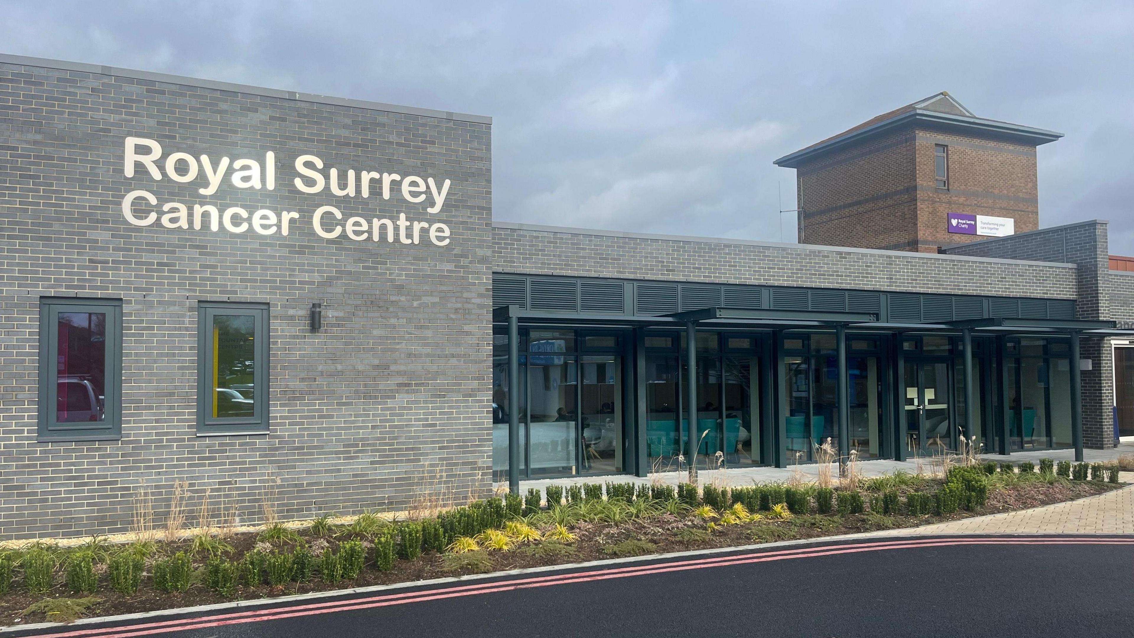 The front of the Royal Surrey cancer centre, it is a grey building with silver lettering. There are plants along the front. 