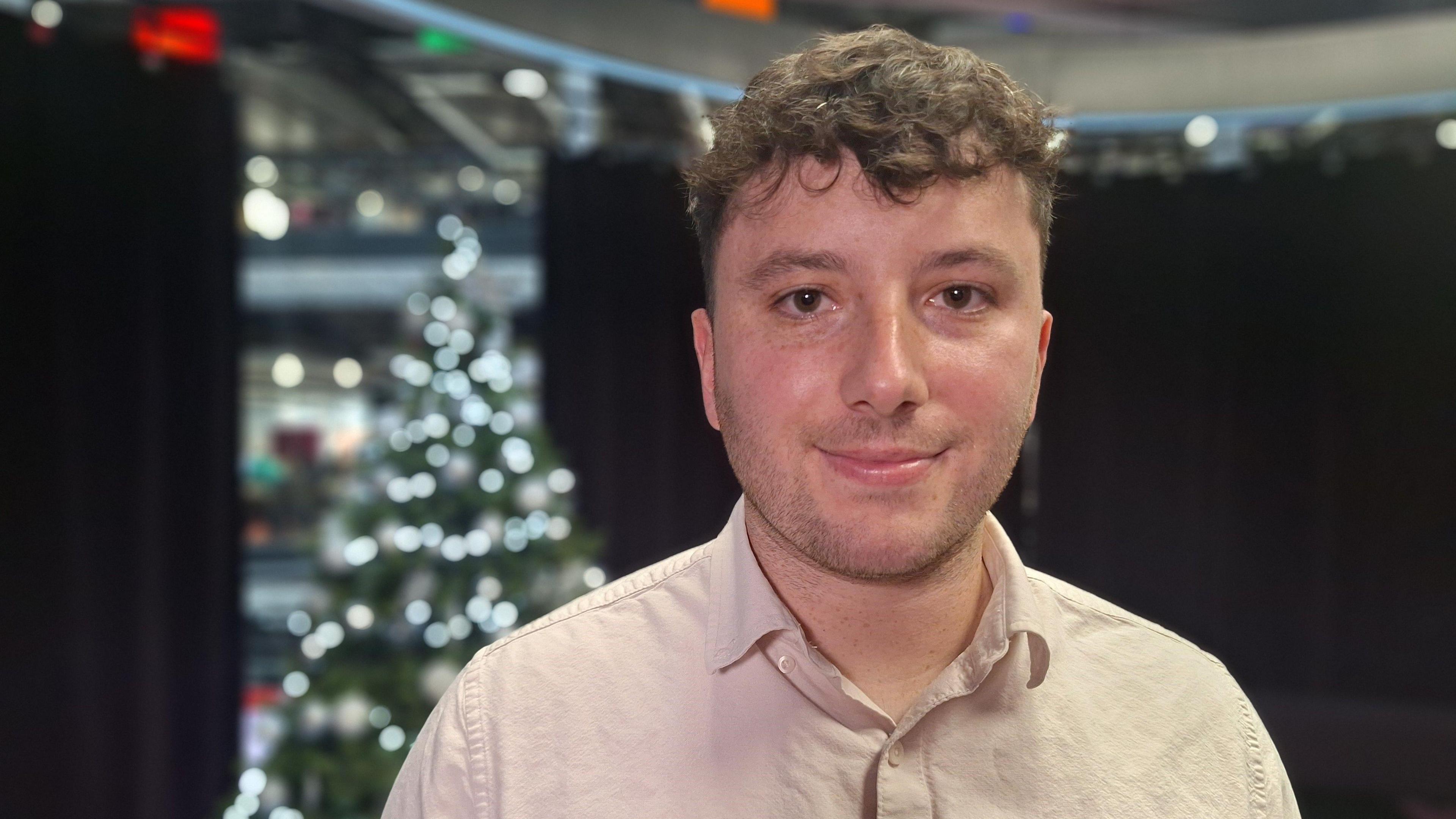 Dyn ifanc gyda gwallt cyrliog â chrys golau yn edrych i'r camera. Mae coeden Nadolig i weld yn y pellter tu ôl iddo.