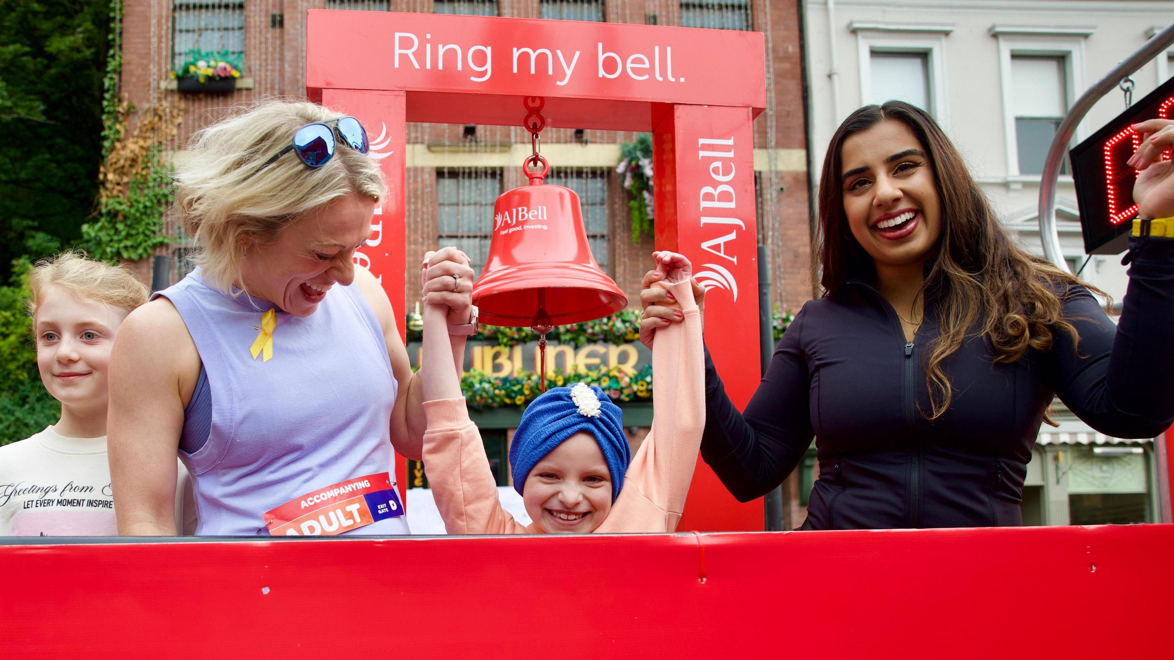 Two women, one with blonde hair and a purple vest, and the other with long dark hair and a black zipped long sleeved t-shirt, standing either side and holding the raised arms of a smiling six-year-old girl wearing a blue turban-style head-dress. Behind them is a large red bell hangs from a red stand.