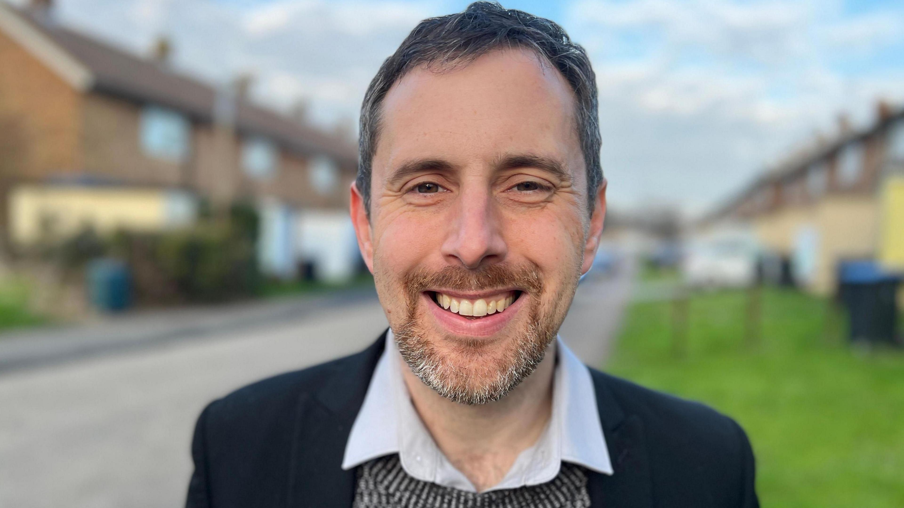 Chris Vince smiling while standing on a residential street. He has short black hair and stubble. He is wearing a black blazer with a knitted grey jumper and white shirt underneath.