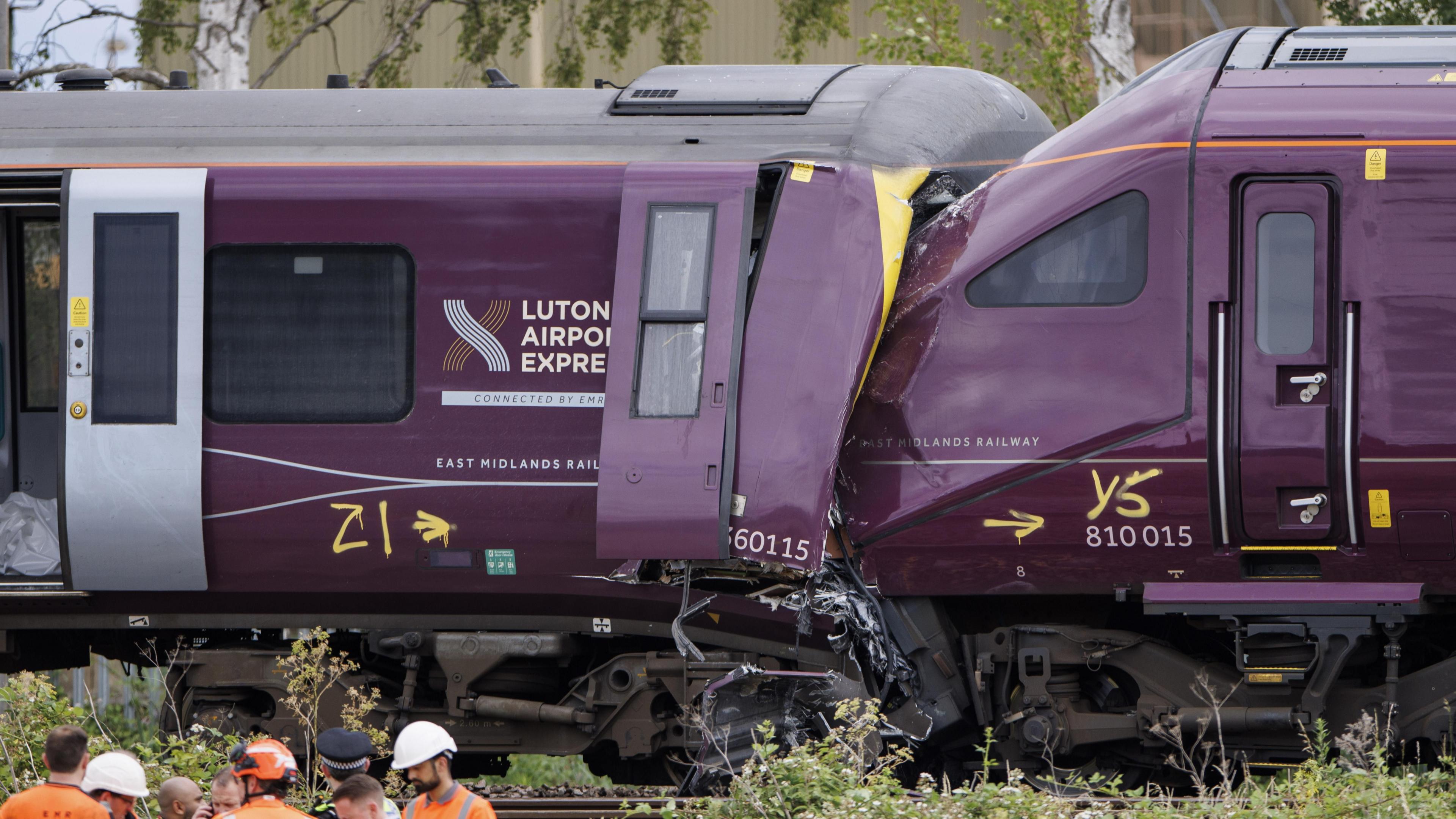 Two damaged trains, the front of one purple train is crushed into the end of another, with severe impact damage visible around the cab area.