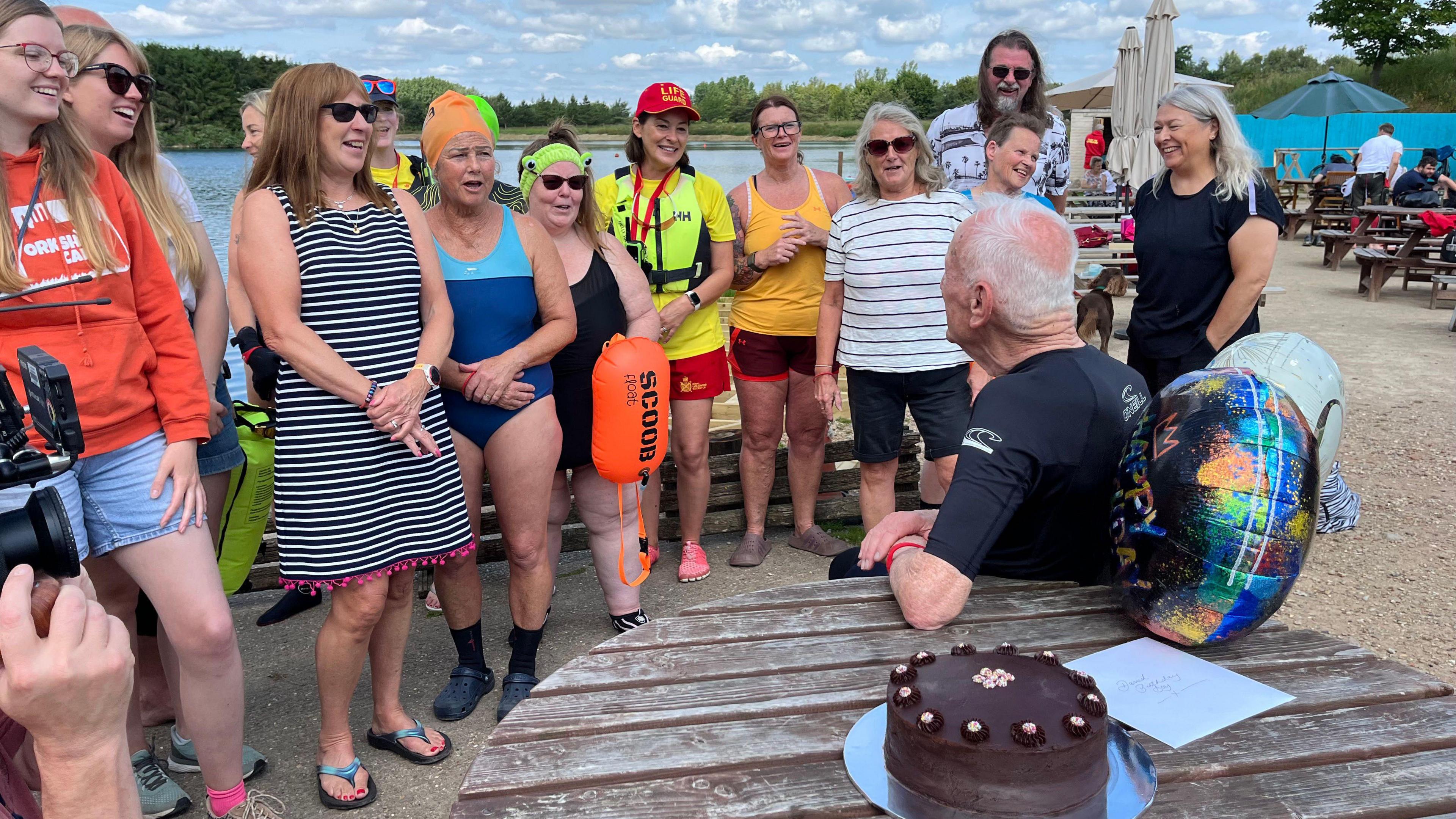 Swimmers and a lifeguard stand around as David sits in the centre with a cake and balloons