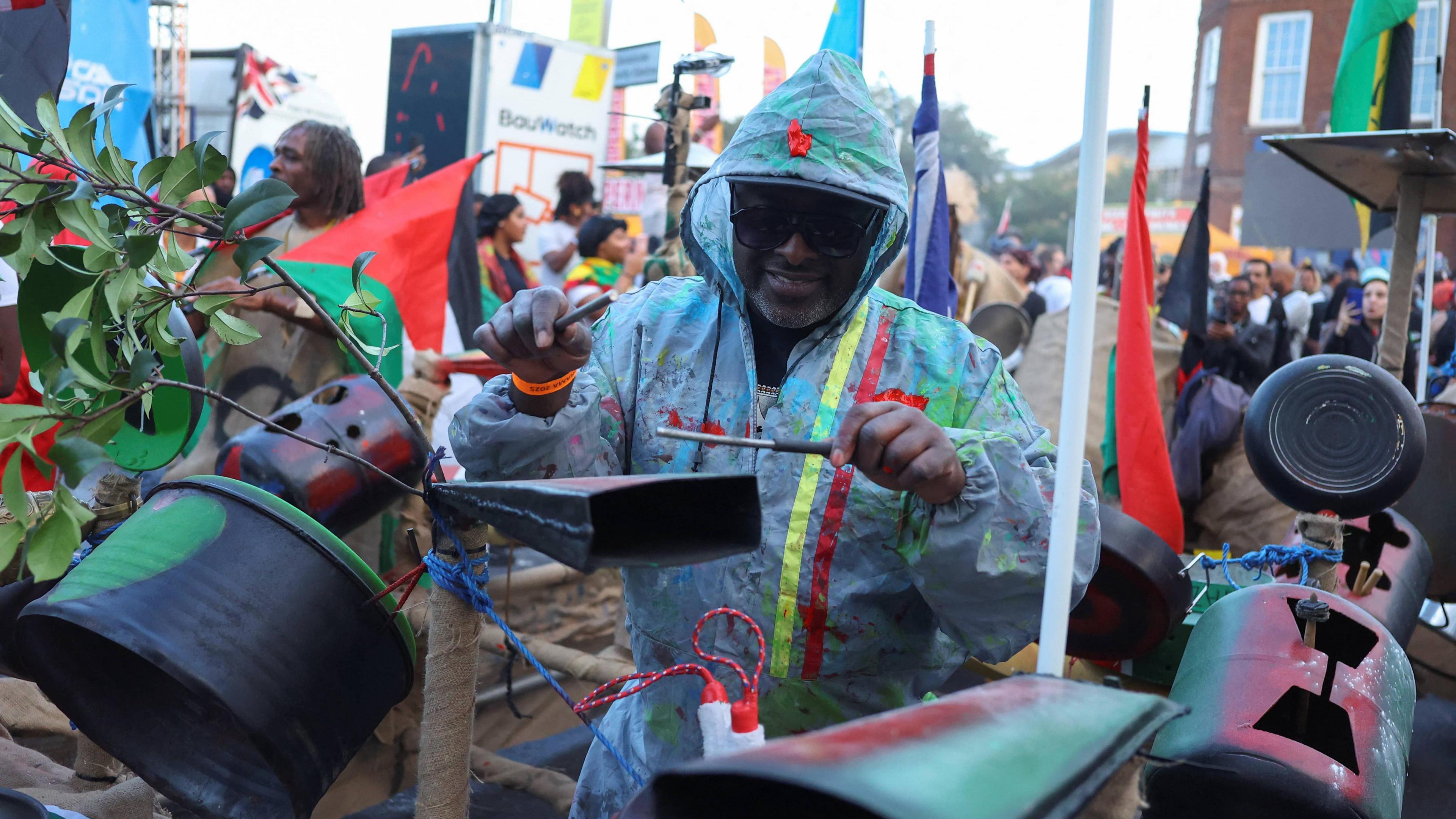 A man in plastic overalls covered in paint plays music on a metal cowbell. Crowds of people take part in the celebrations behind him. 