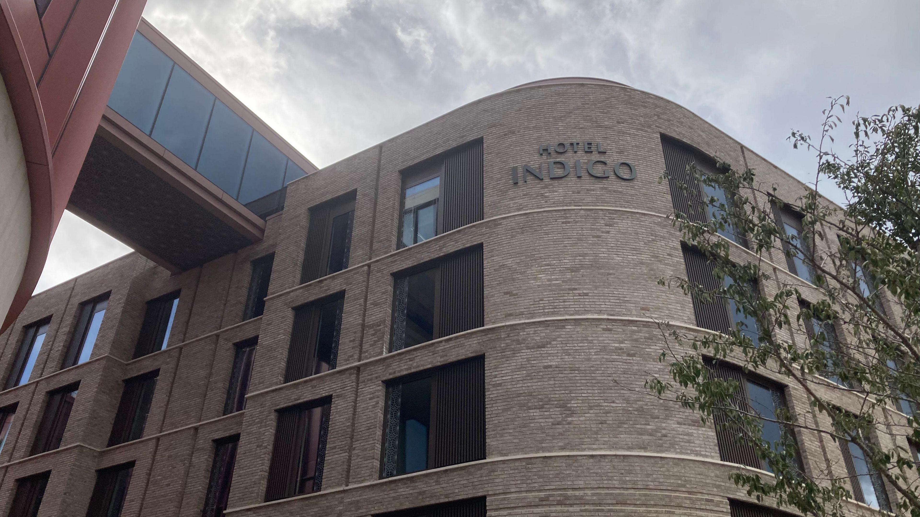 A tall brown brick building has a curved corner, upon which is the name Hotel Indigo. A large glass bridge connects the top of the hotel to the neighbouring office building.