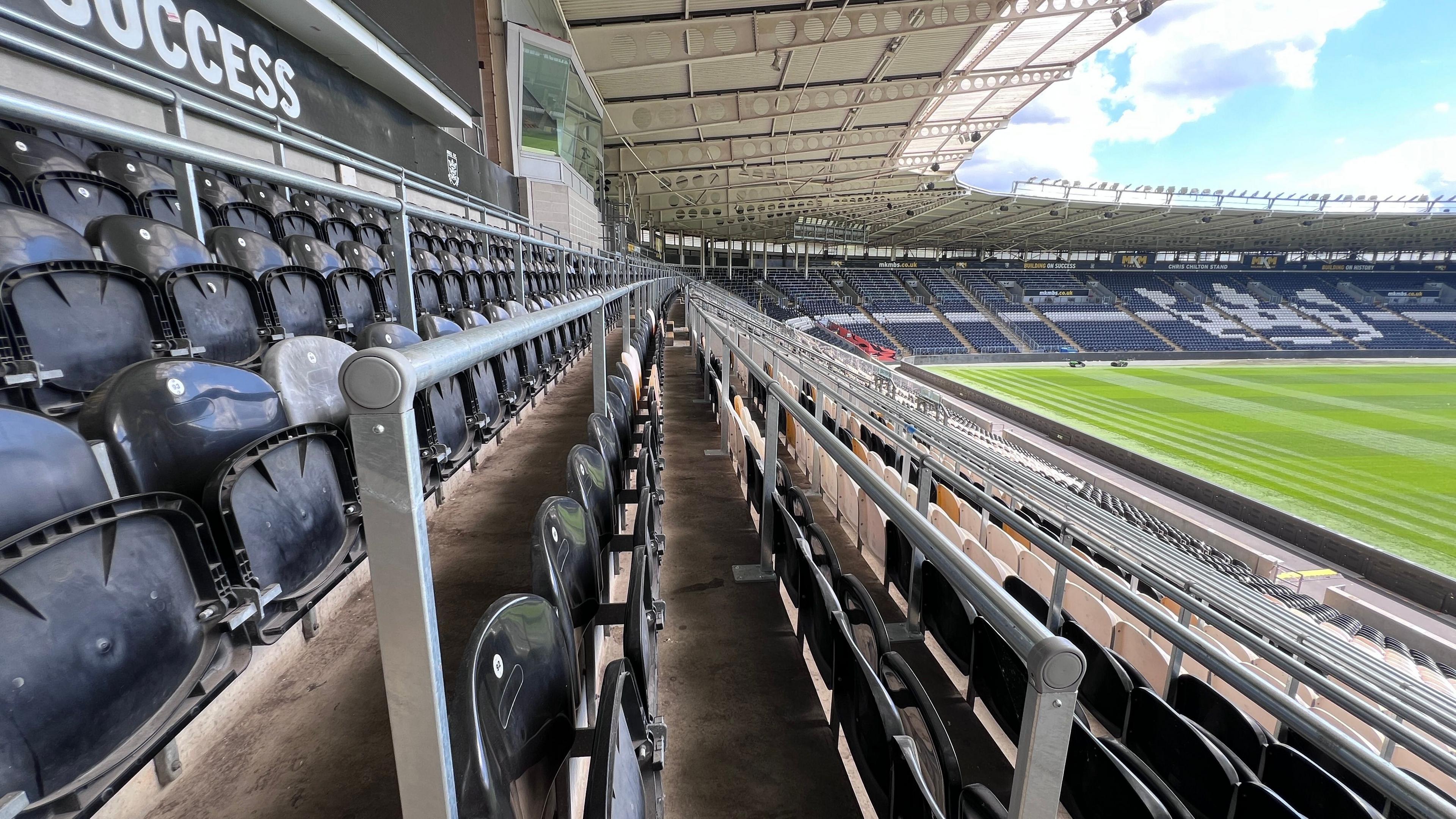 The view along a row of safe standing seats at a sports stadium. Round steel barriers extend into the distance, each in front of a row of black plastic seats. The grass pitch is visible to the right.