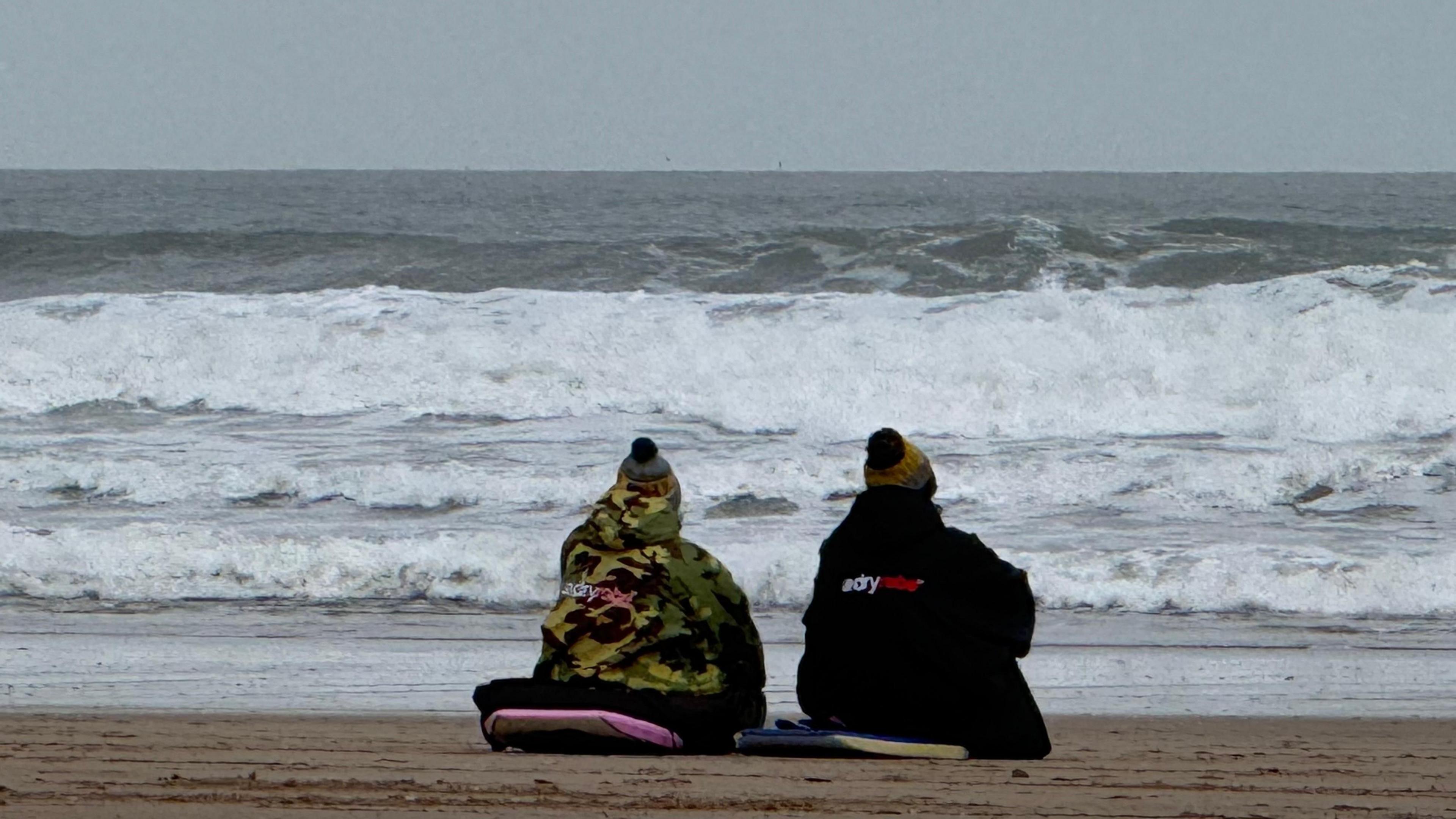 Two people wearing Dryrobe coats sit on surfboards at Croyde beach. They have their backs to the camera as the waves crash into the beach on a grey day.