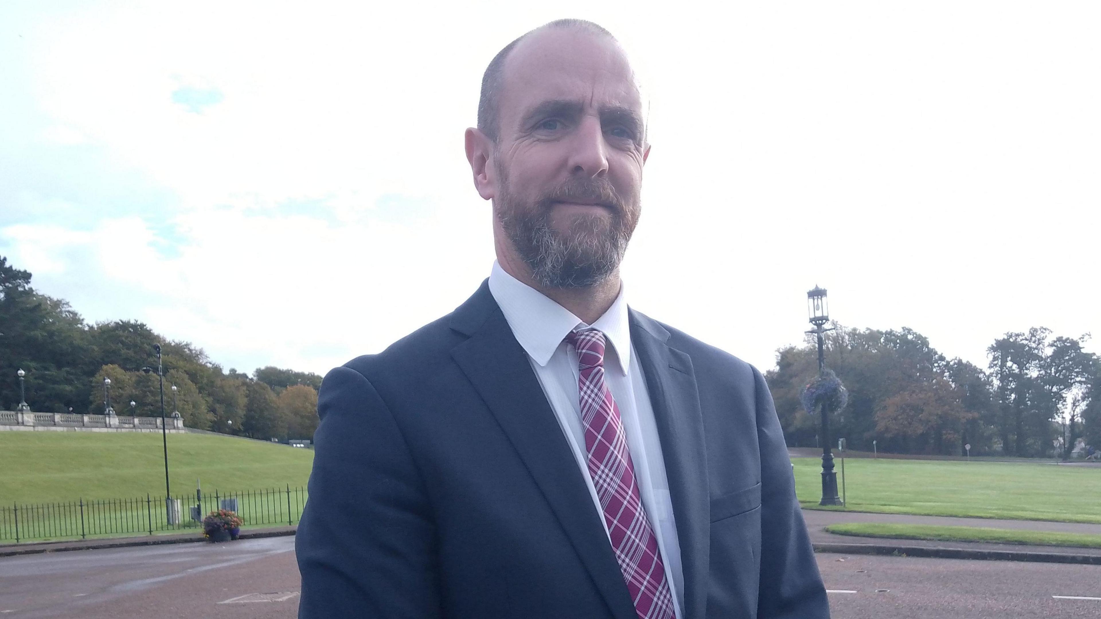 Mark H Durkan, who is bald, wears a black suit, white shirt and red tartan tie. 