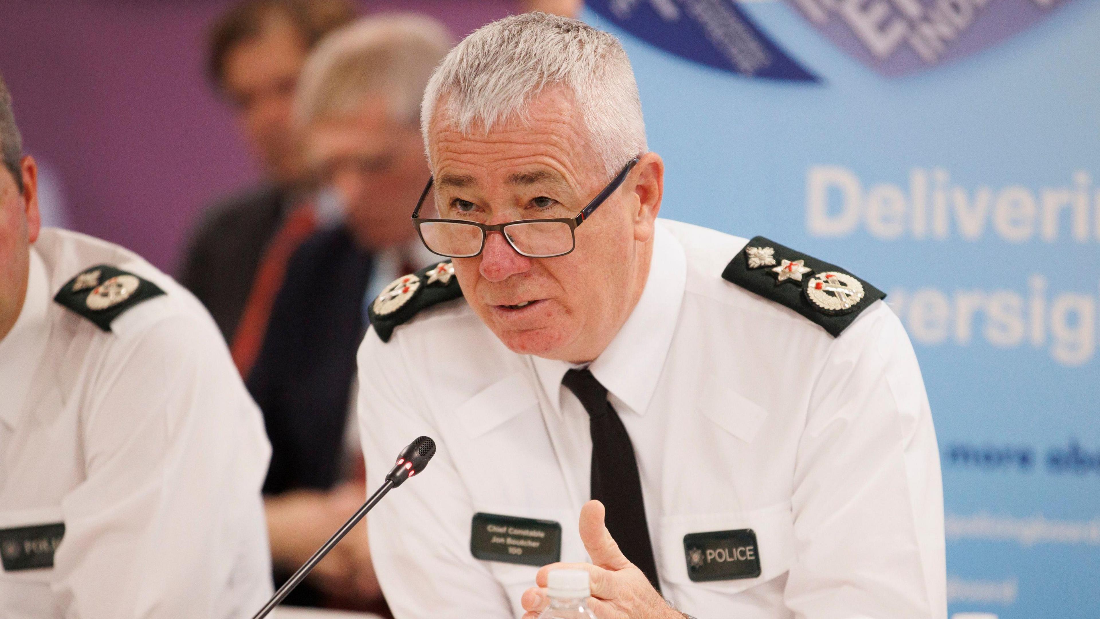 Jon Boutcher - a grey haired man wearing black square-framed glasses and a police uniform sits in front of a microphone.