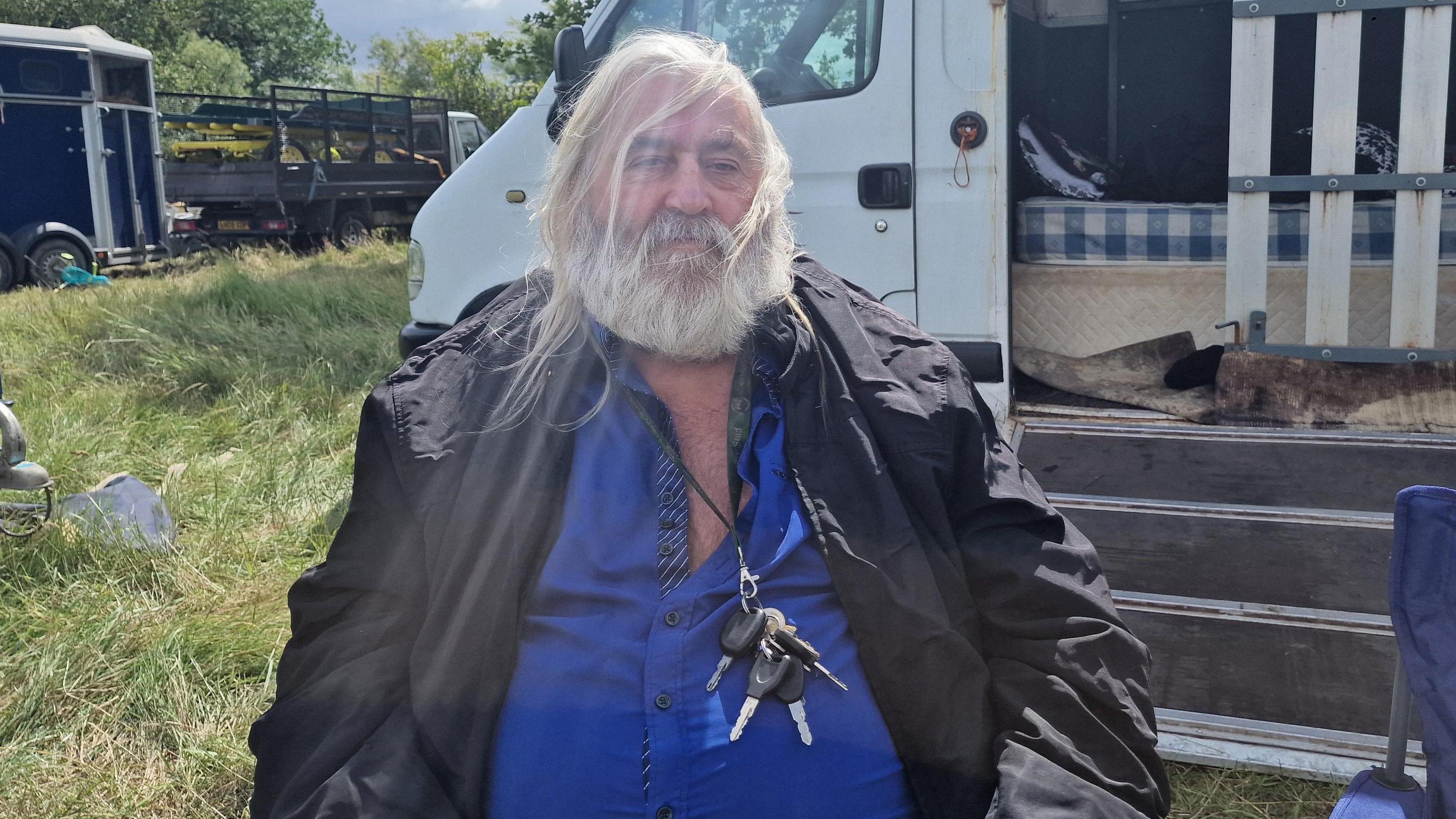Man with long white hair, a black jacket and blue top sat in front of a white vehicle. There is grass behind him.