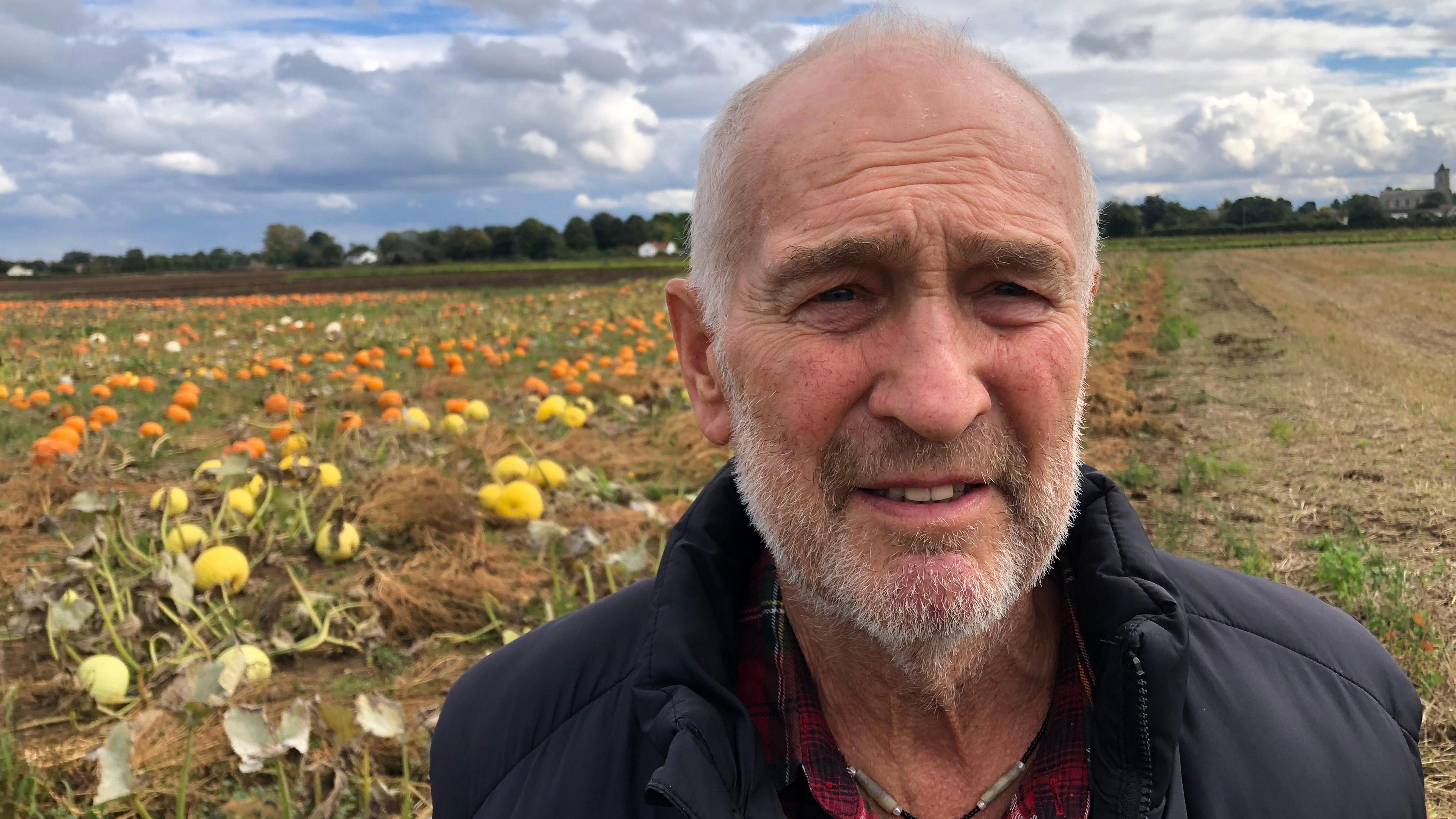 John Quince, 71, has thinning grey hair with matching beard and moustache. He is standing in front of a field of pumpkins at his farm in Isleham. He is wearing a red-checked shirt and a black body warmer.