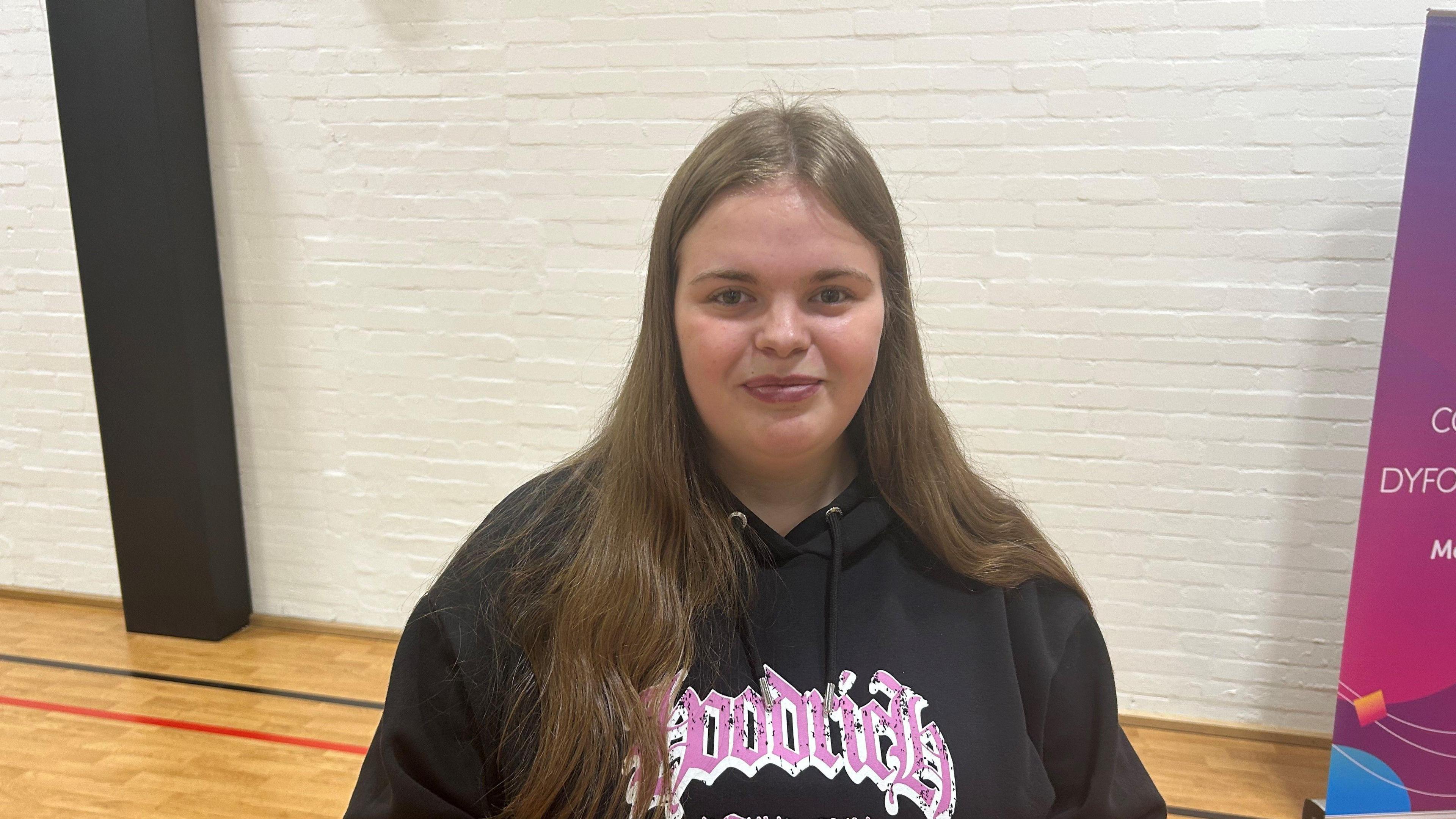 Ruby smiling at the camera, she is wearing a black jumper with pink and white lettering. She has long brown hair.