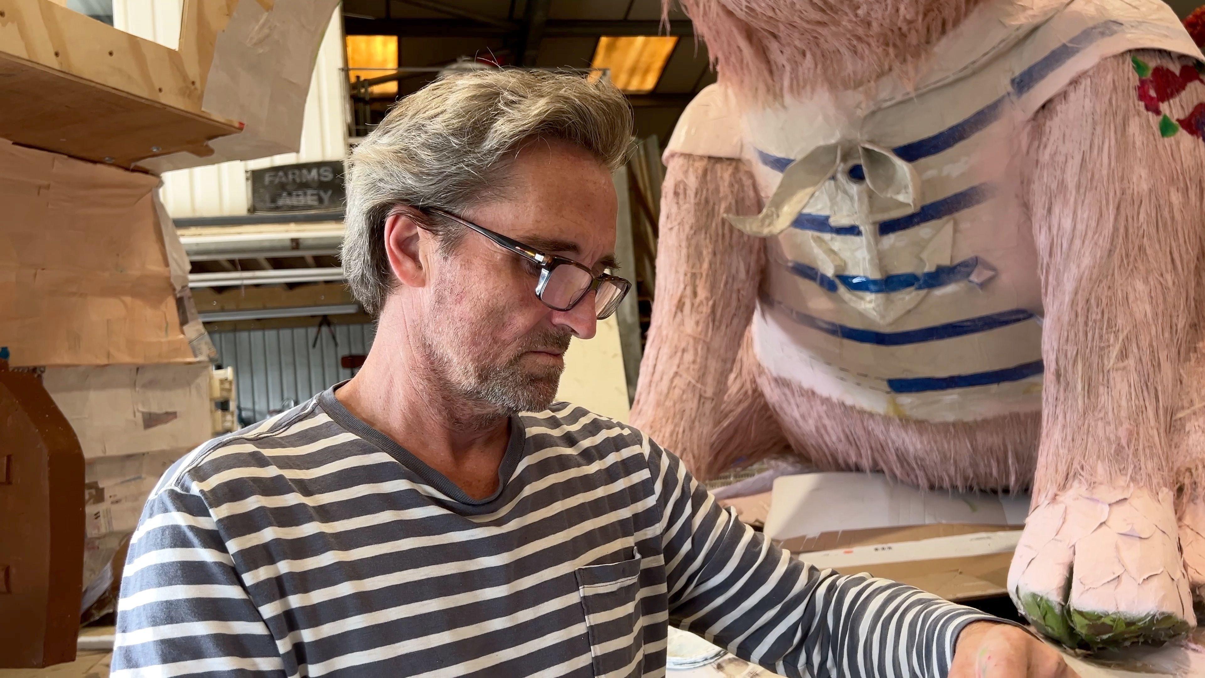 Marcus Davies with glasses on in front of the St Ouen float. Behind him is the body of a giant paper mache pig. He's wearing a striped top and looking down