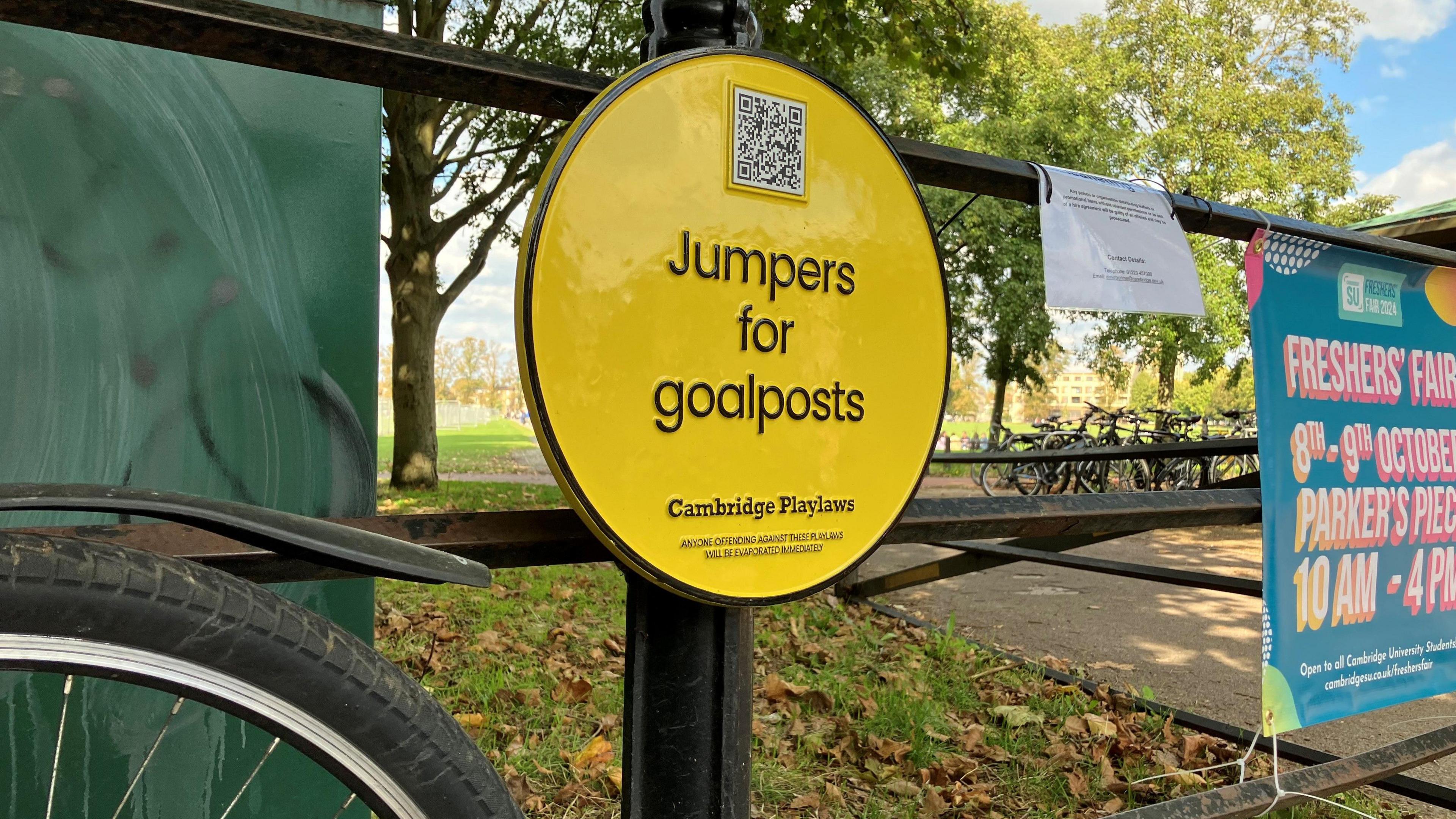 A yellow sign says 'Jumpers for goalposts' and 'Cambridge Playlaws'. The sign is on a metal railing and bikes can be seen in the background