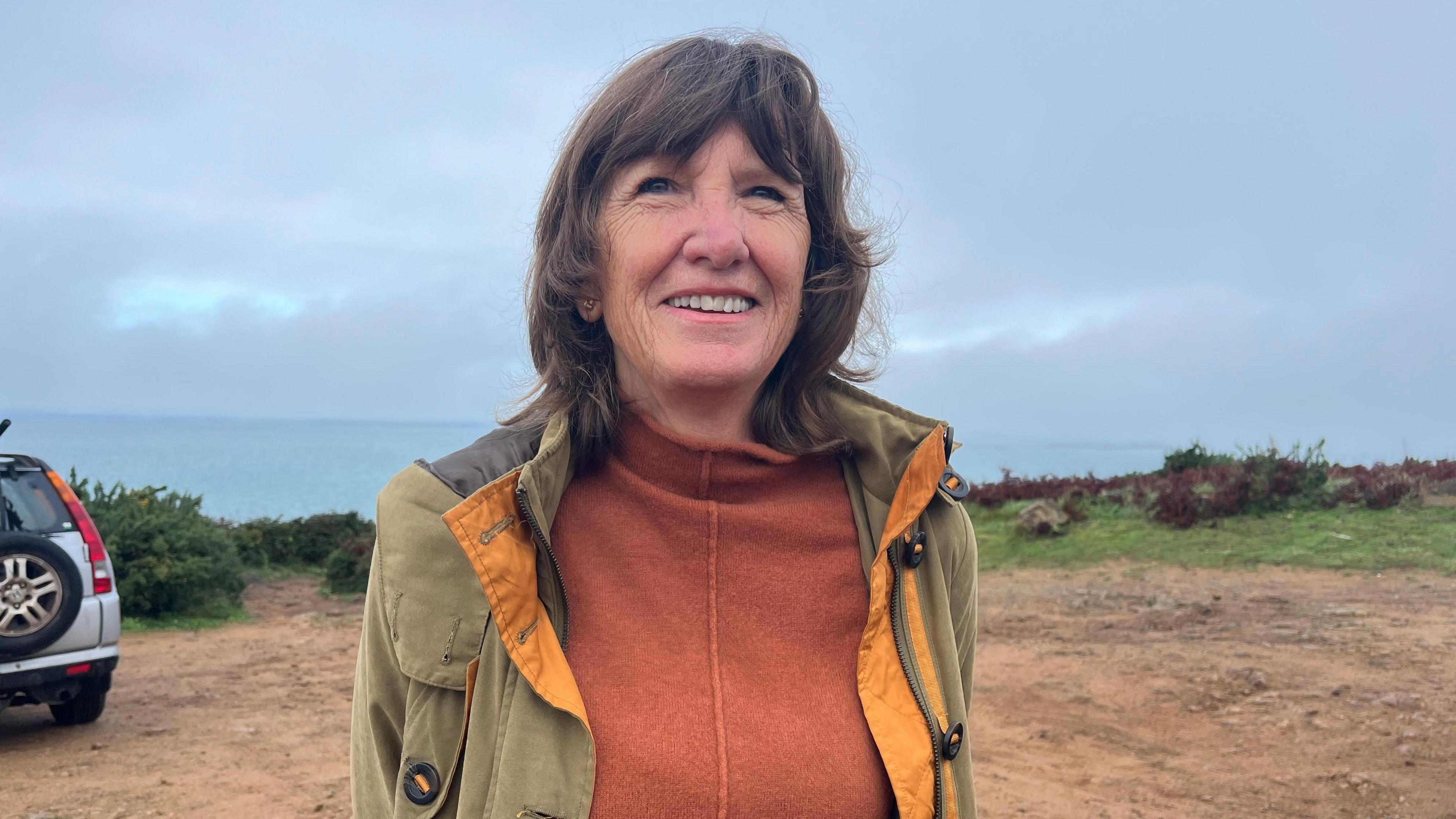 Nicolette Westwood - a woman with dark brown hair and a fringe smiling, wearing an orange knitted jumper with a khaki coat over it. She is standing in a ground car park with the sea and grey skies behind.