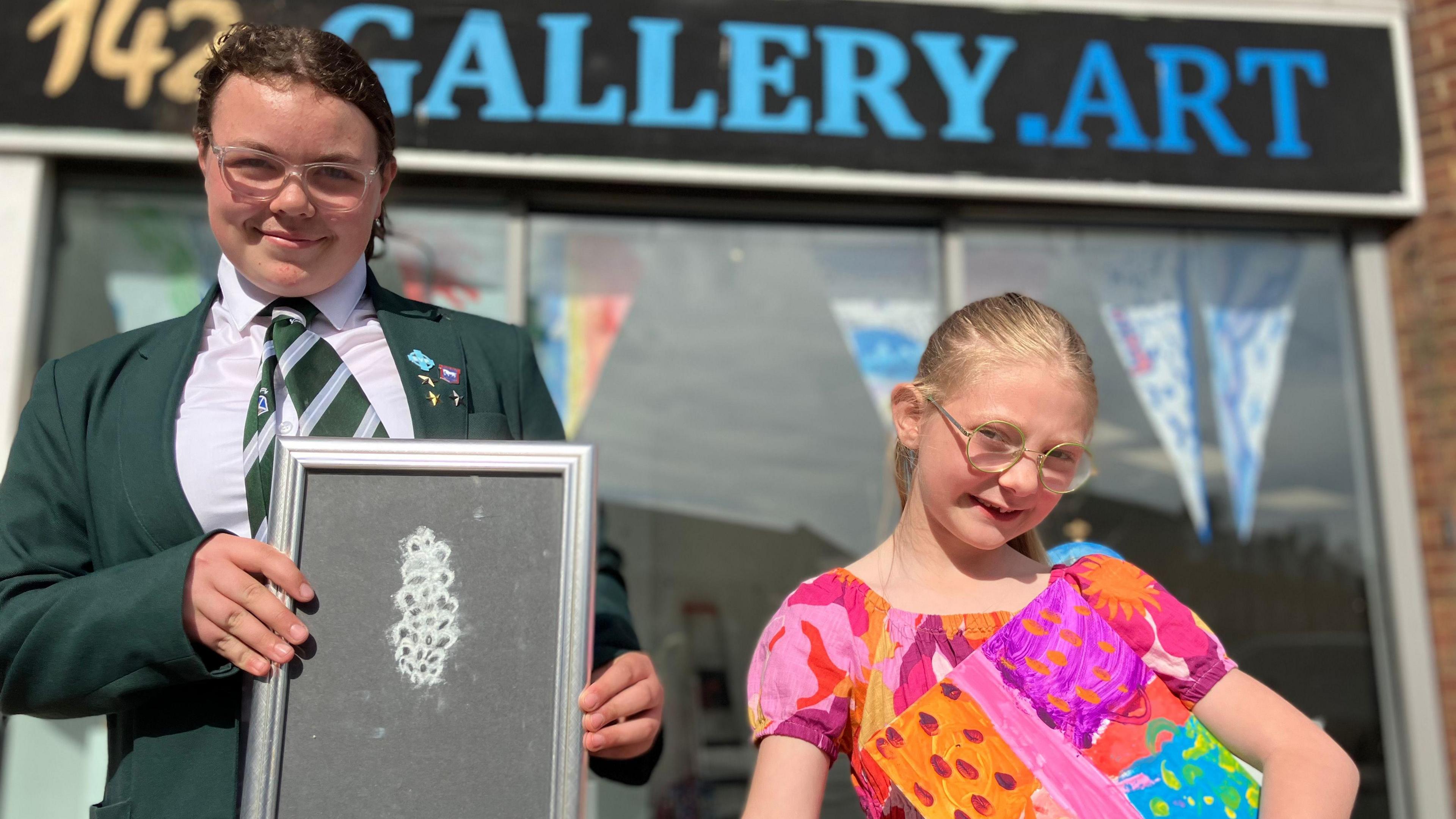 Two young people stand in front of a shop window. The sign at the top reads "142 gallery.art". They are both holding up pictures, one is a white chalk design on black paper and the other is brightly coloured squares in pink and orange