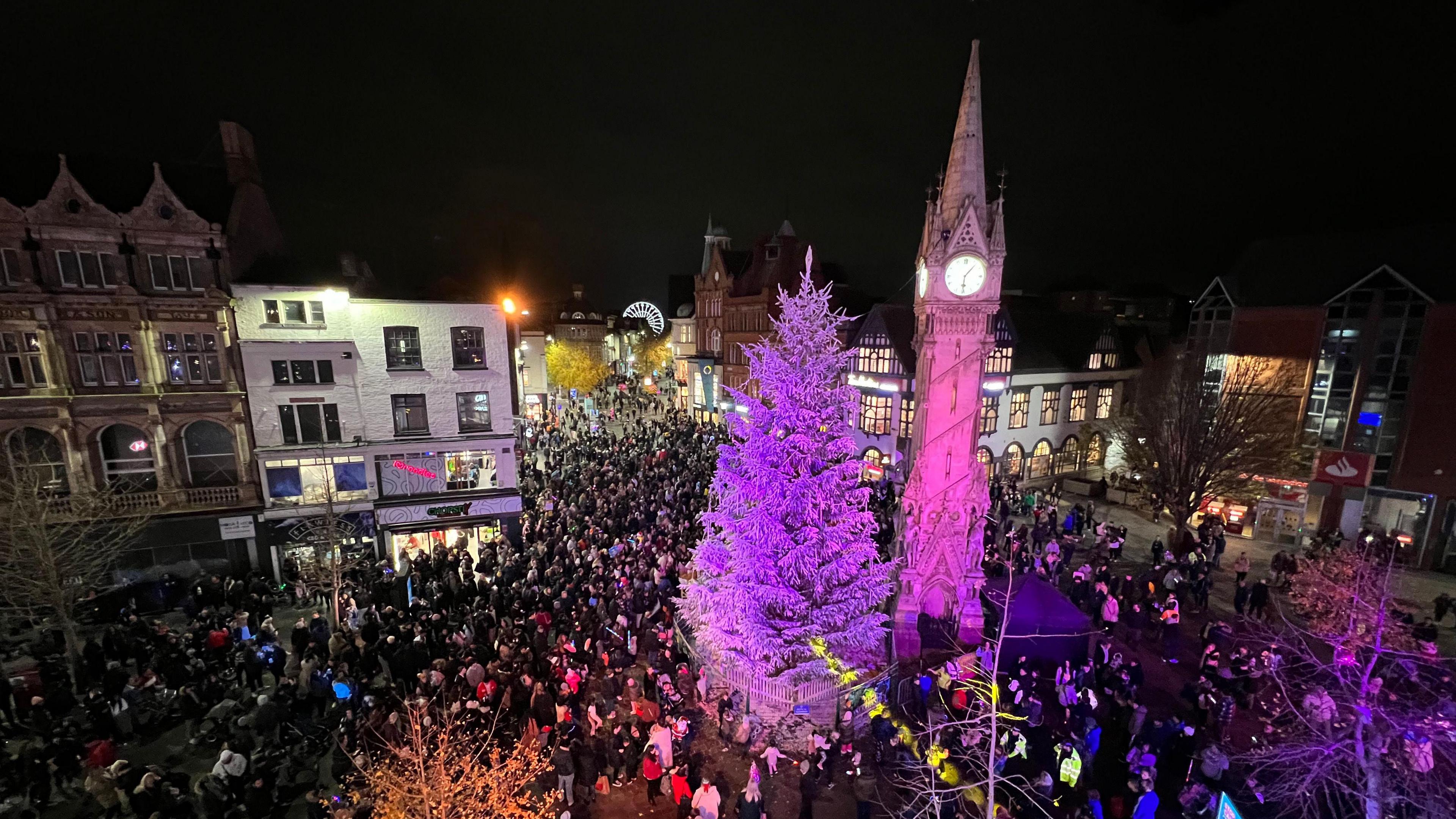 A view from above the crowds gathered around the Christmas tree and Clock Tower in the centre of Leicester at night, with colourful lighting illuminating the features