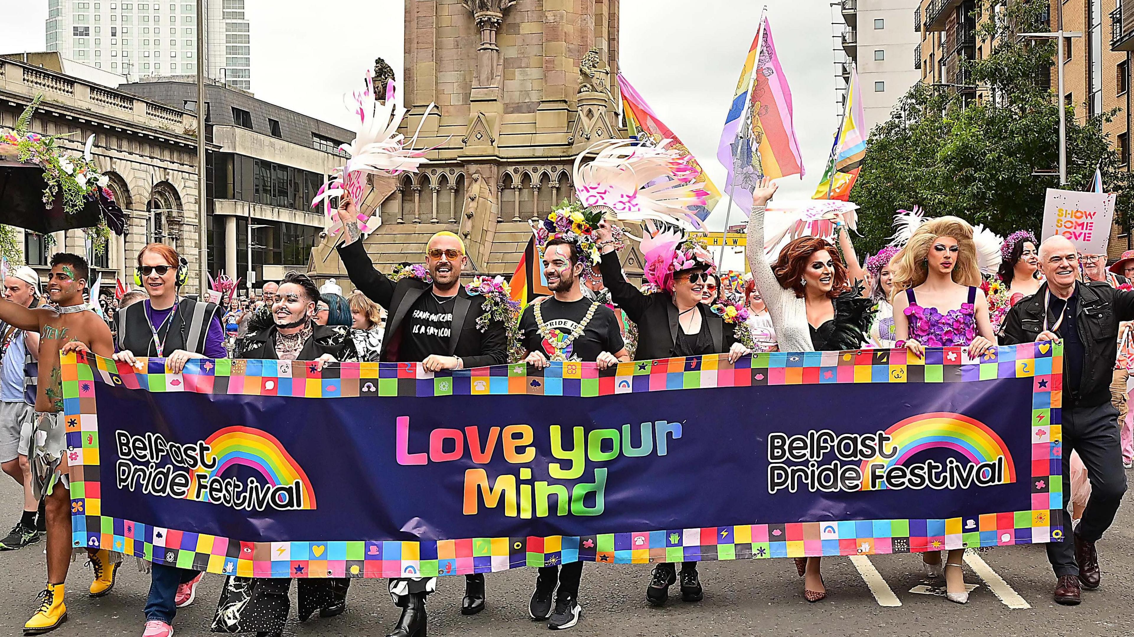 A group of people marching through the road. They are smiling. Some are waving feathers, posters or flags. The banner they're holding says Love Your Mind. Belfast Pride Festival. The Albert Clock is behind them.