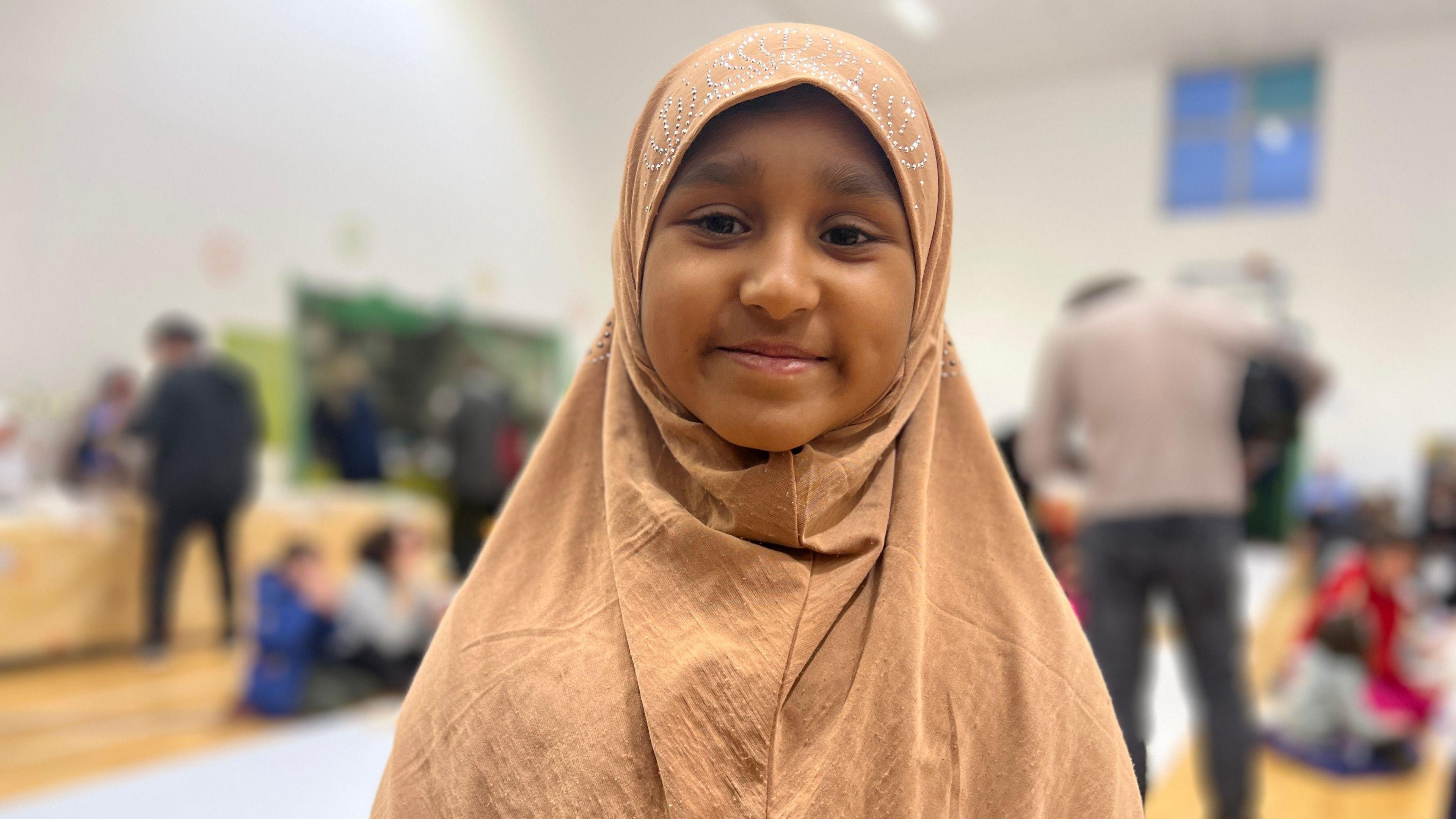 Salwa, a young girl in a peachy-brown headscarf, smiles into the camera.