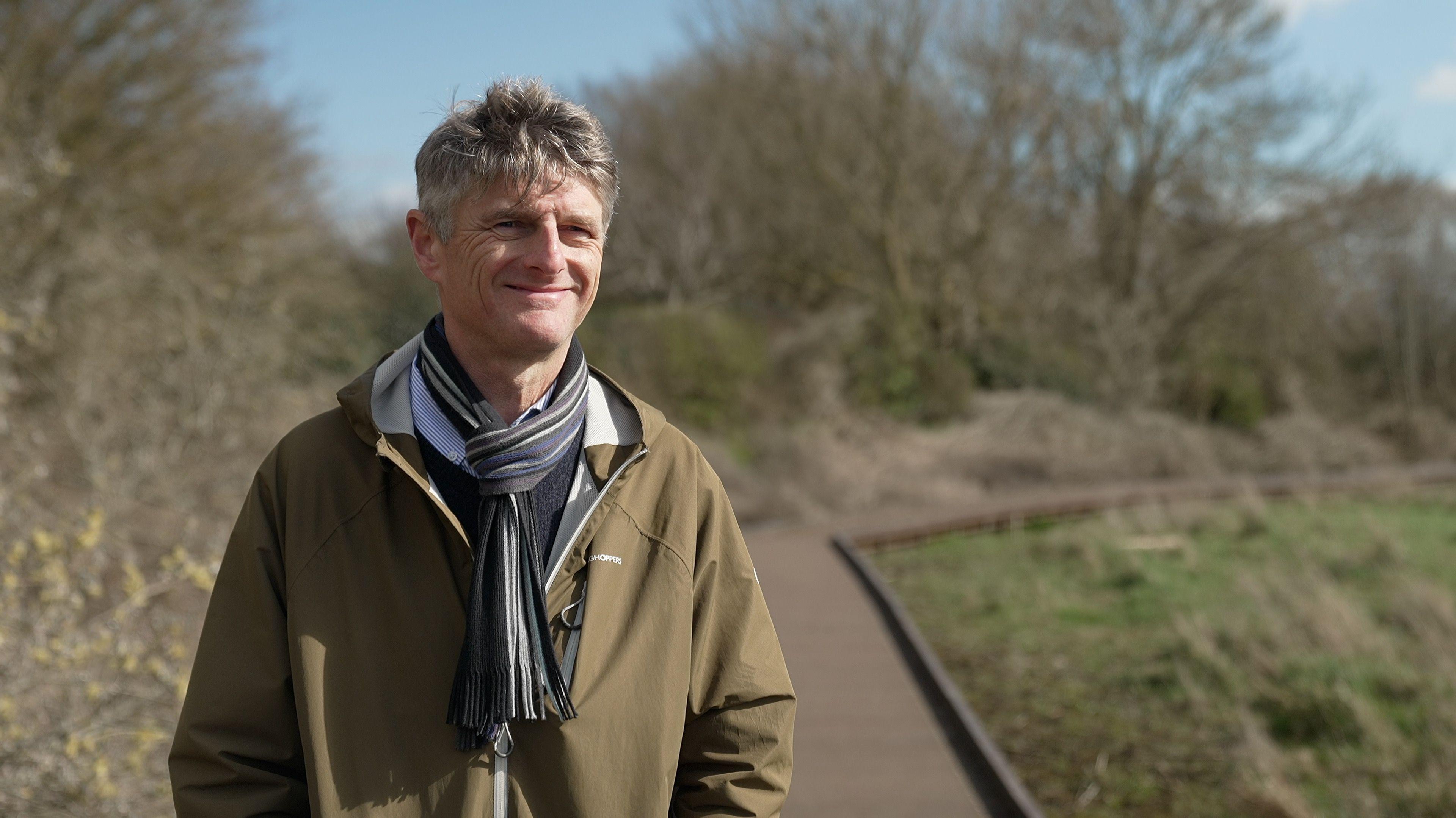 A man with silver hair and a stripey scarf weaing a khaki jacket is standing with a smile on a wooden boardwalk. Trees in the background have no leaves and the piece of land next to the walkway has some green grass but is mostly mud.