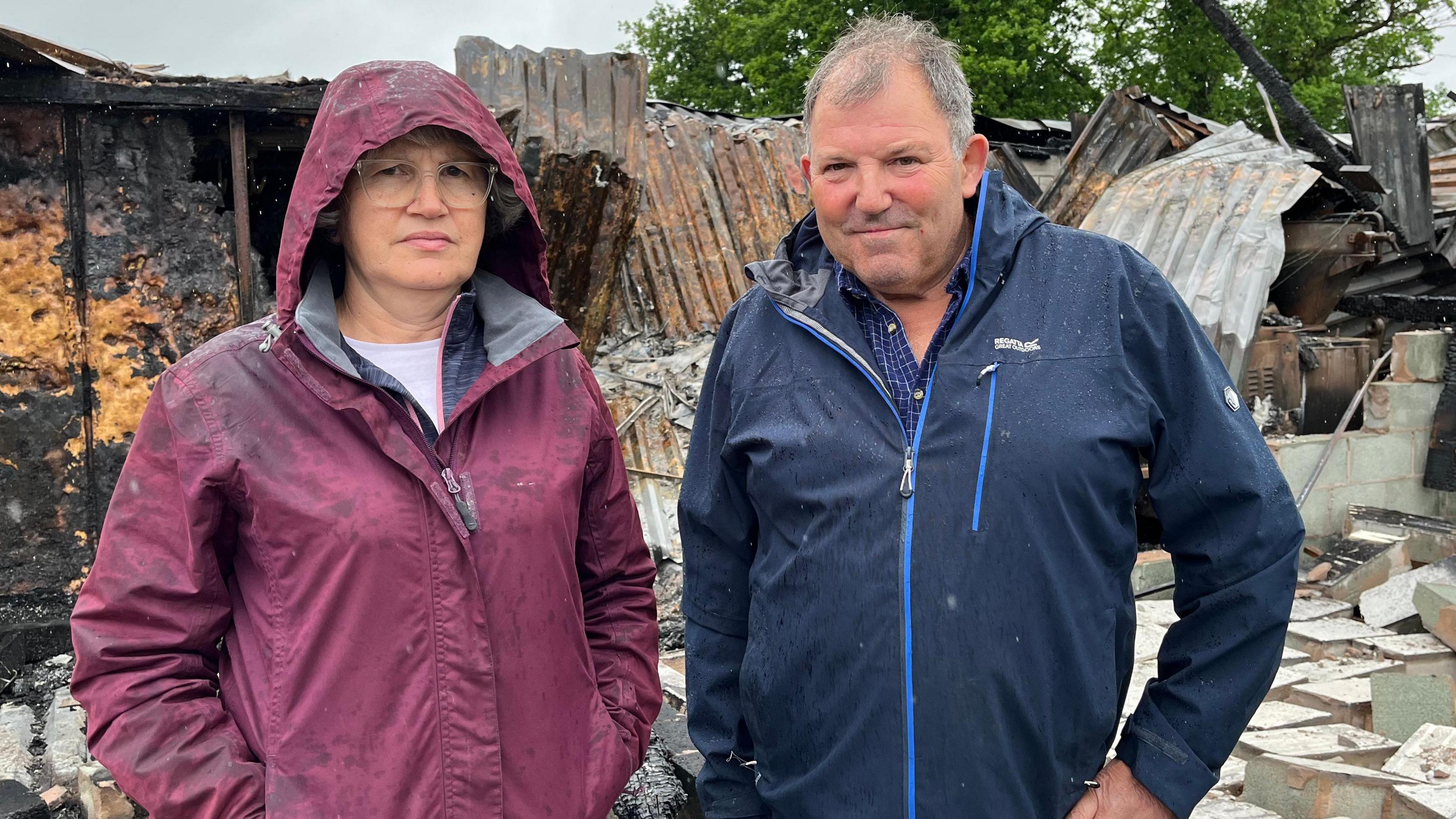 A woman is wearing a red coat with her hood up and a man is wearing a blue rain jacket. They are standing in front of a burnt building and rubble.