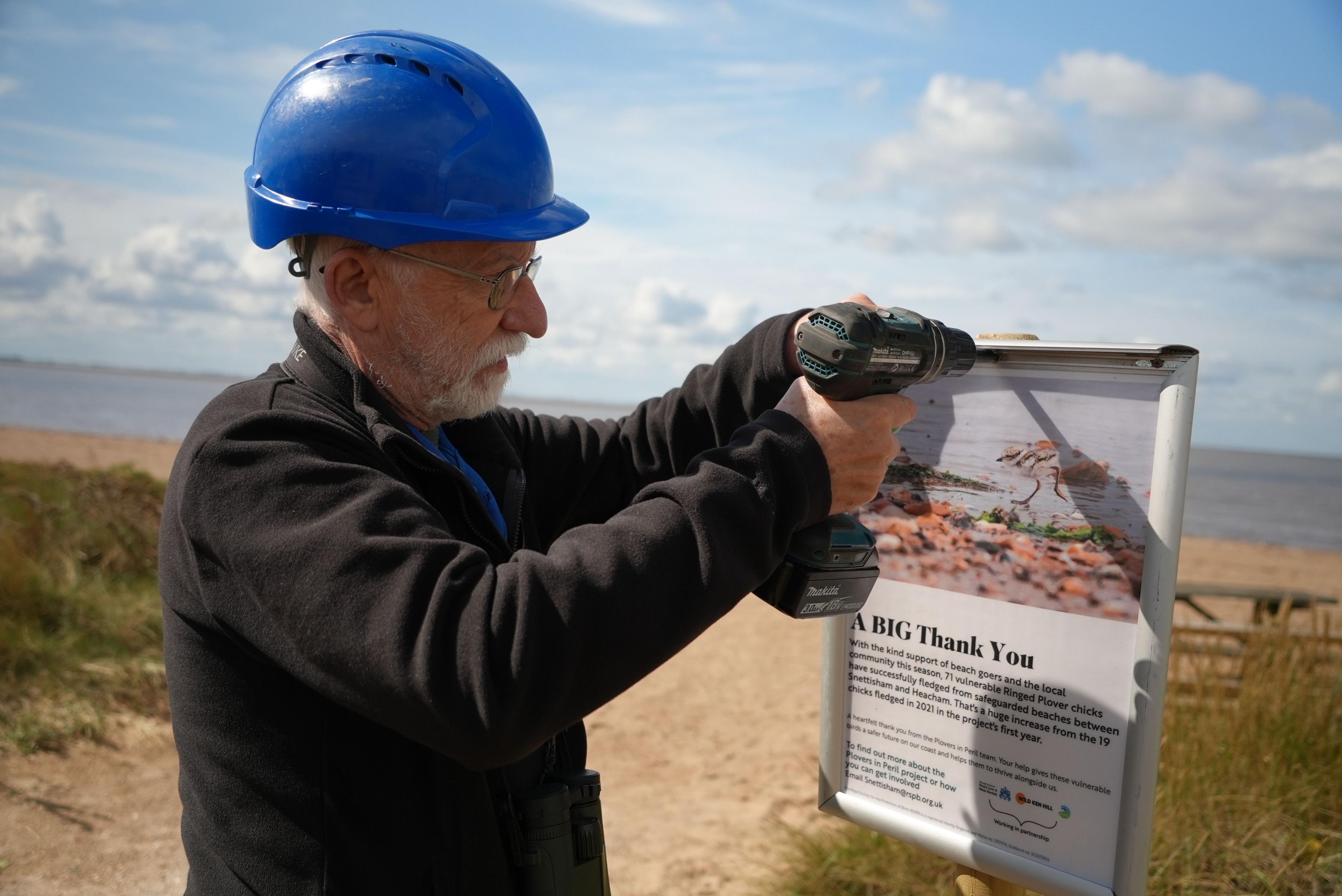 Man wearing a hard hat with a drill. He is putting up a poster on wooden post on a beach. 