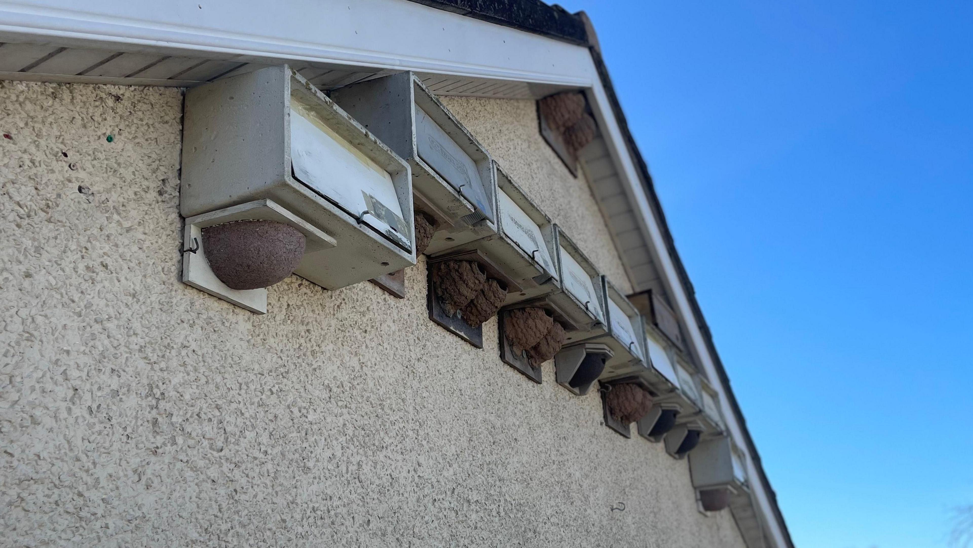 11 white swift boxes and house martin nests sit on the upper side of a stone white wall. They sit just below the arch in the roof, on the side of a house. 