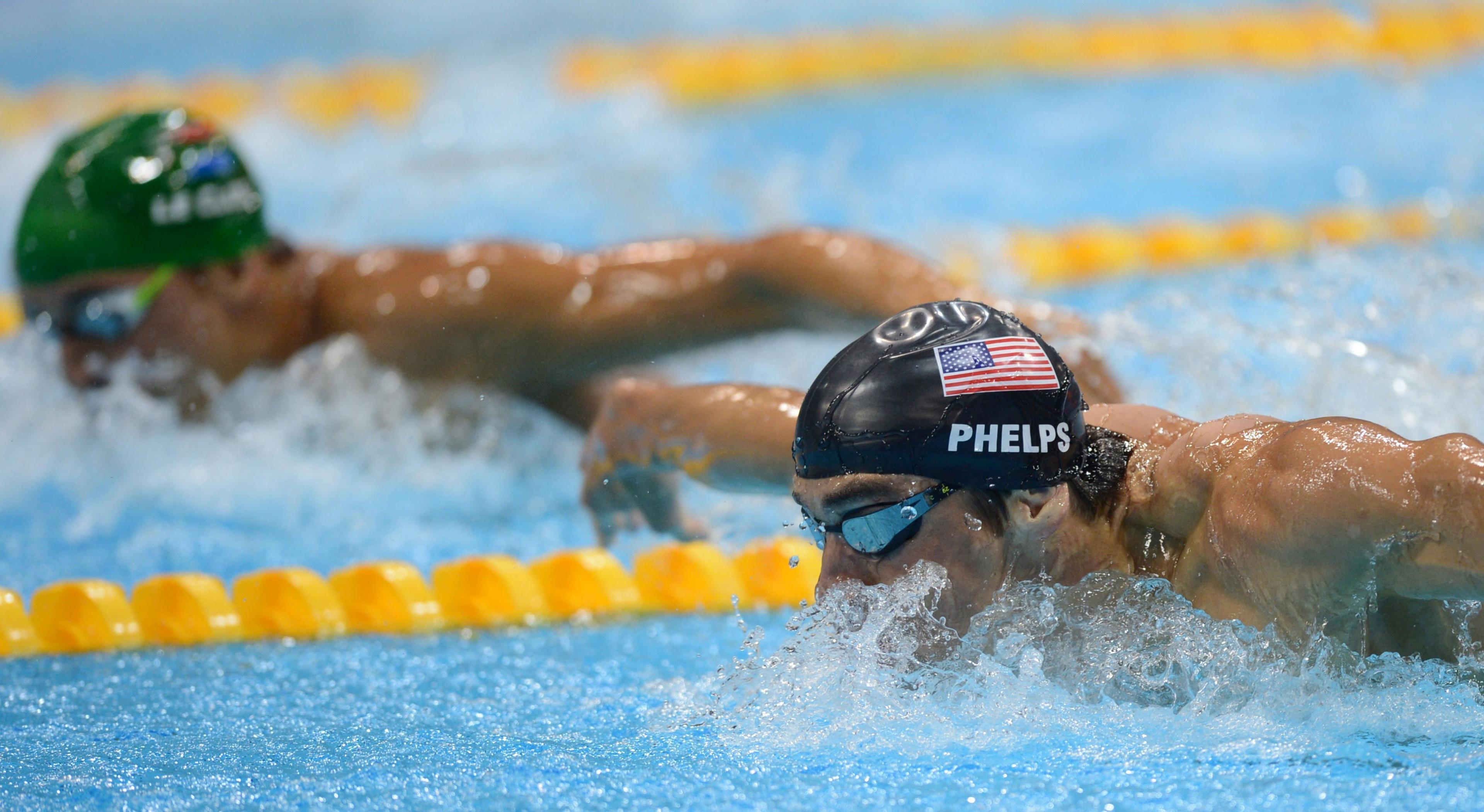 Chad le Clos and Michael Phelps racing in a swimming pool. They are both wearing googles. Le Clos is wearing a green swimming cap and Phelps is wearing a black one.
