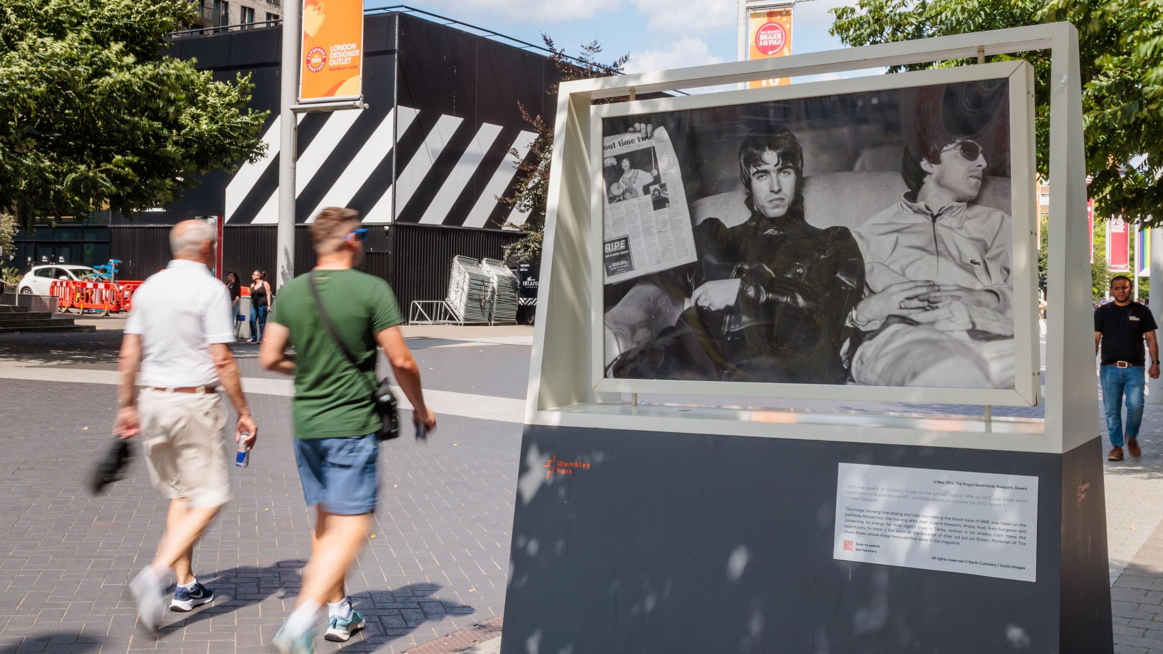 two pedestrians walking past another picture in the exhibition