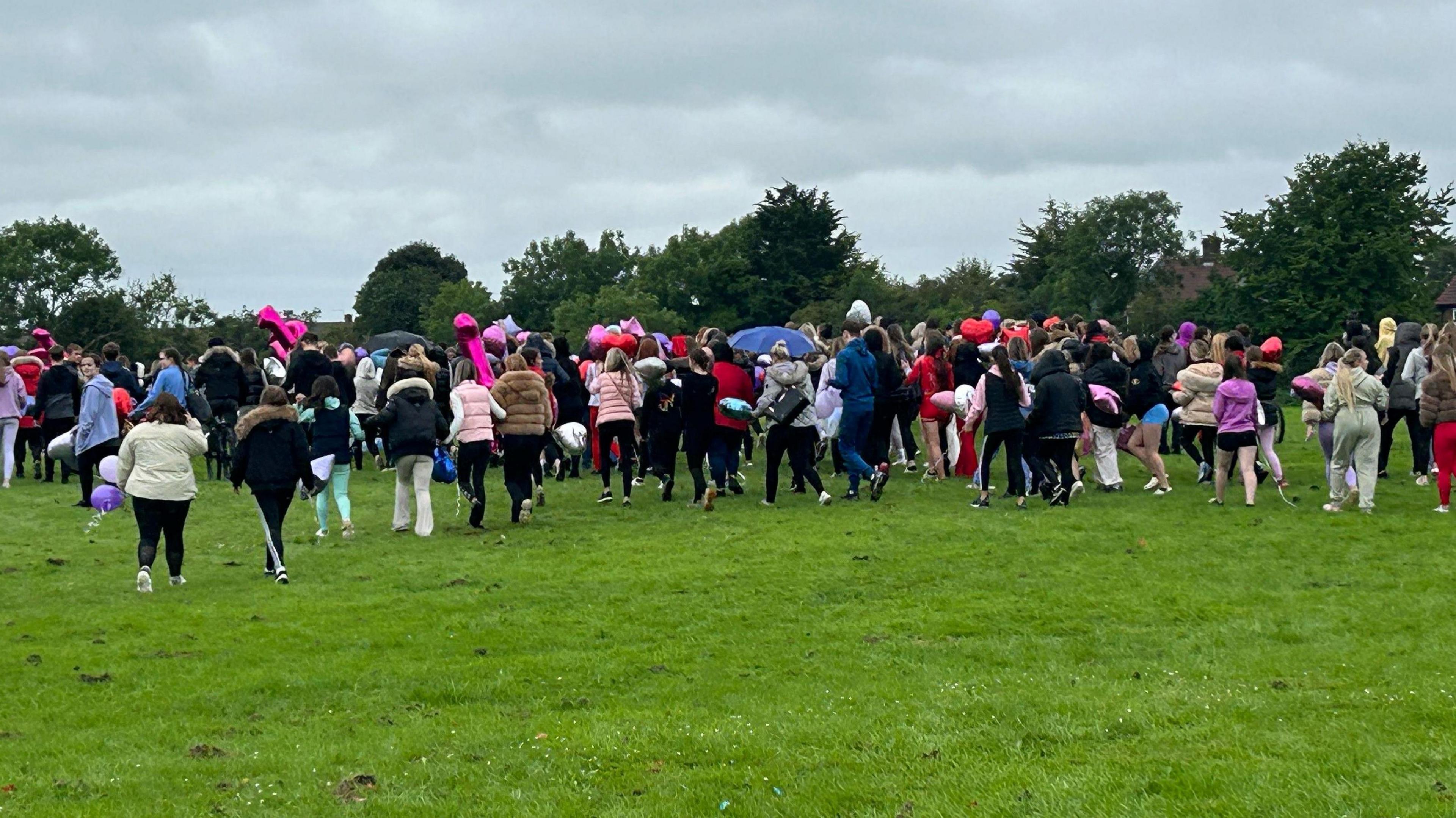 Pictured from the back, crowds of young people gathered in a park, some carrying balloons