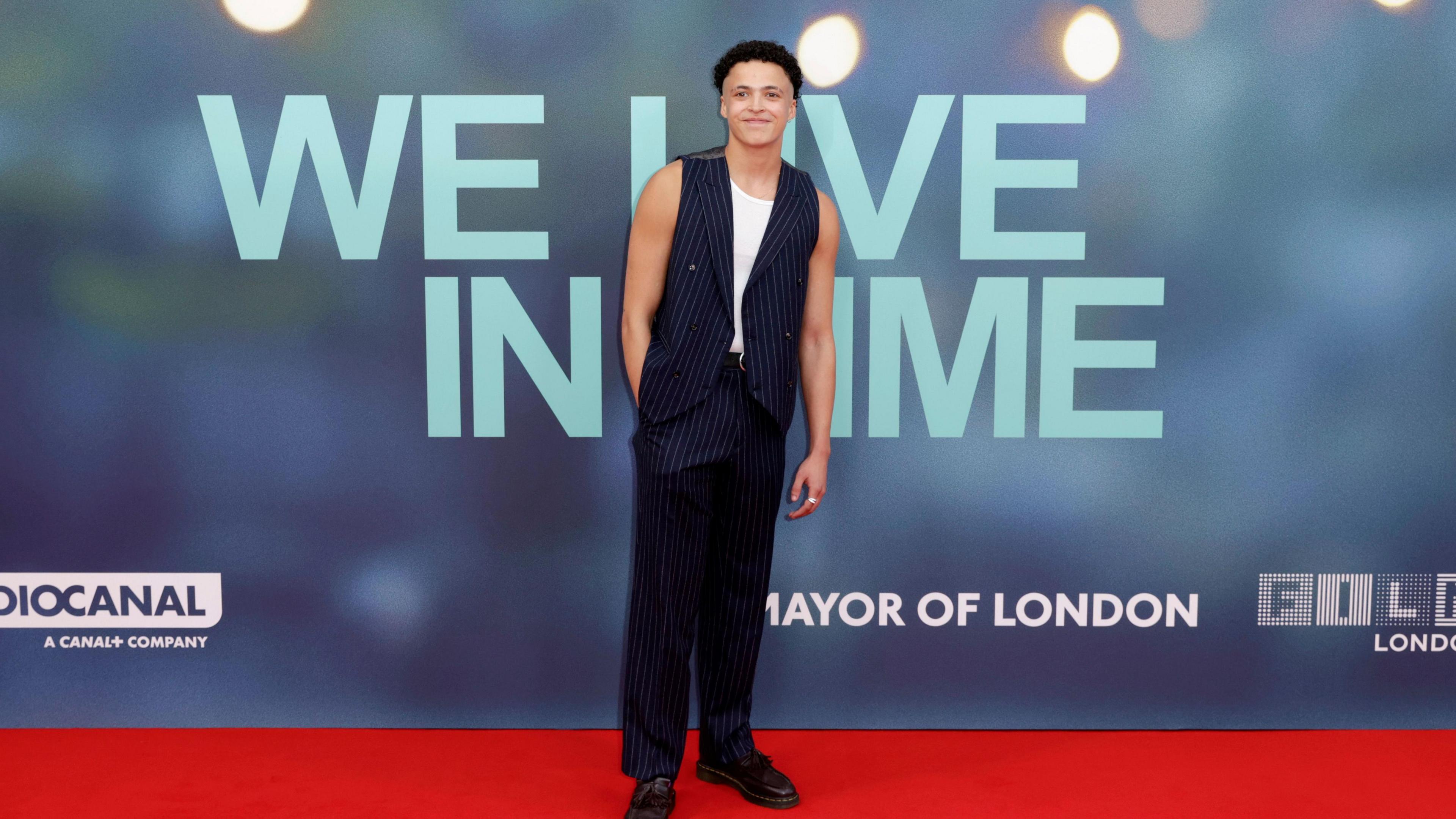 Lee Braithewaite on the red carpet against a blue board that says 'We Live In Time'. They are wearing a dark blue striped waistcoat and matching trousers, with a white vest. They have short, black, curly hair. 