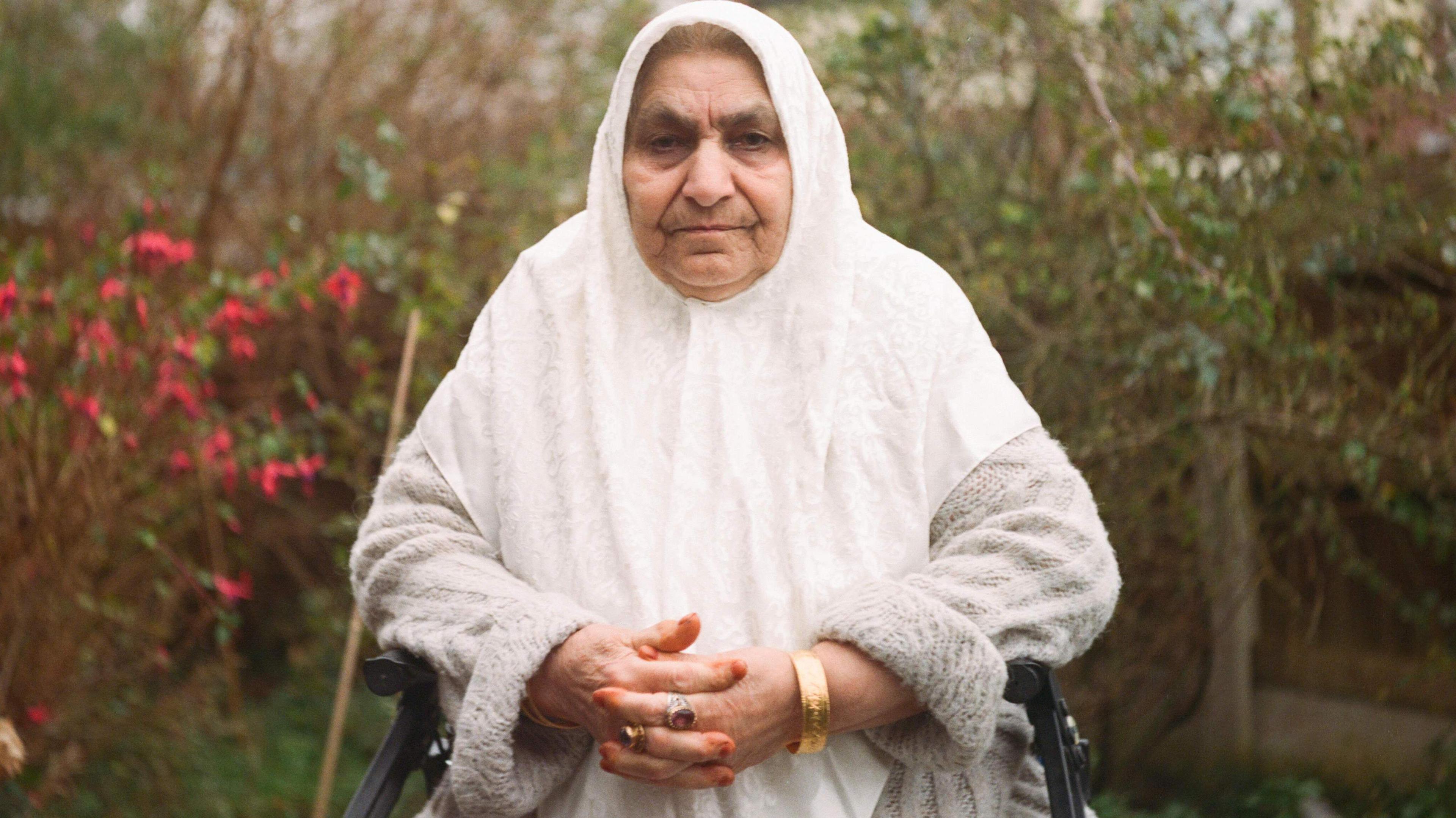An elderly woman wearing a white headscarf and gold bangle sitting in a garden.