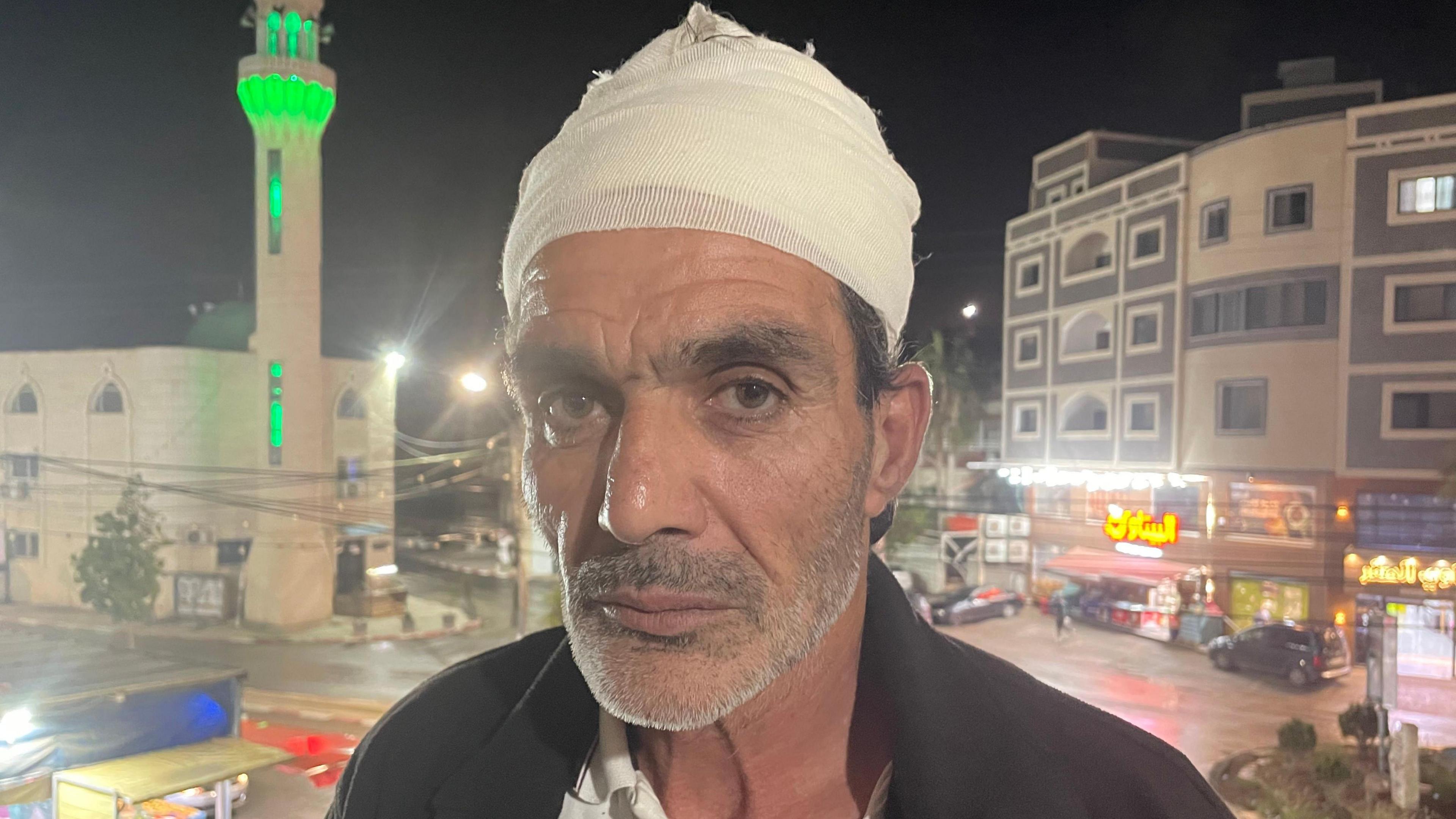 Hamad al-Jagoub abu Rabia. He has a bandage wrapped around his head and a grey stubble beard. Some dark hair can be seen poking out from beneath the bandage and he has thick, dark eyebrows. Behind him are some buildings, including shops and a tower lit up in green.