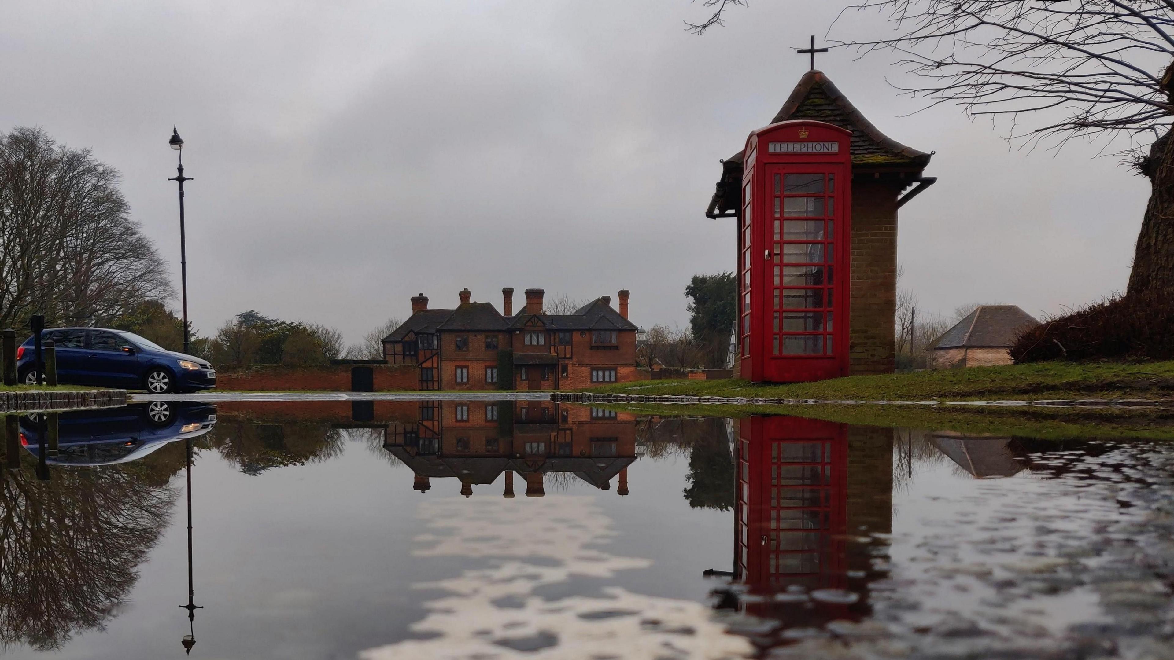 A puddle in the road reflects a red phone box, a brick building and a blue car. The photo is a mirror image of the scene with the water stretching across the road. The cloudy sky is grey. 